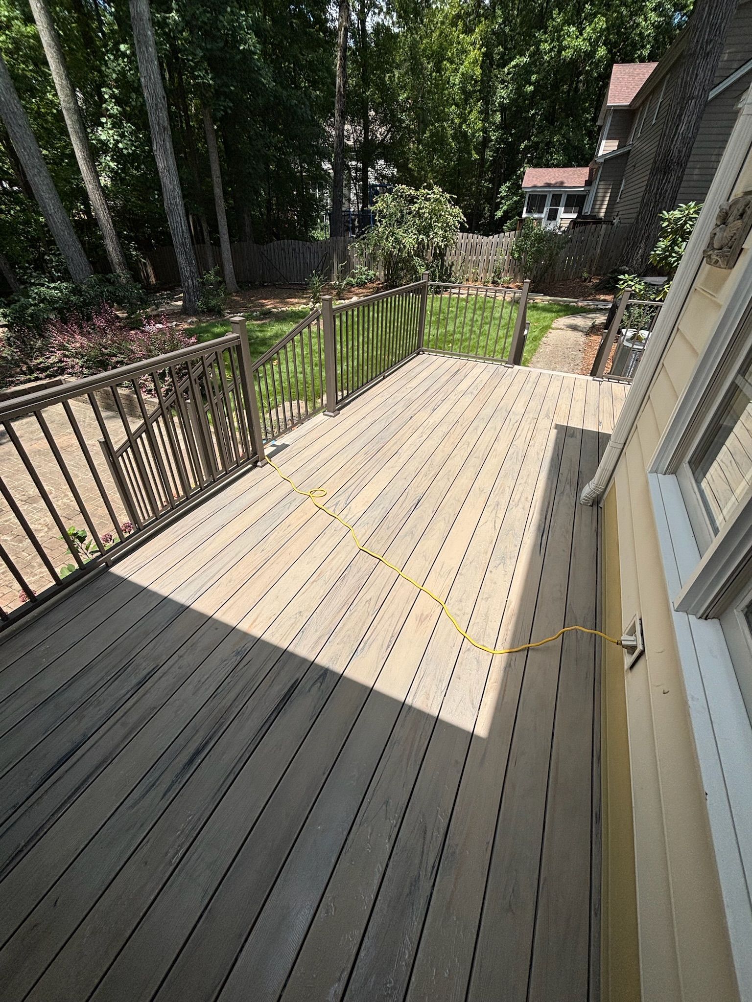 Wooden deck with black railing beside a house, overlooking a green backyard and trees.