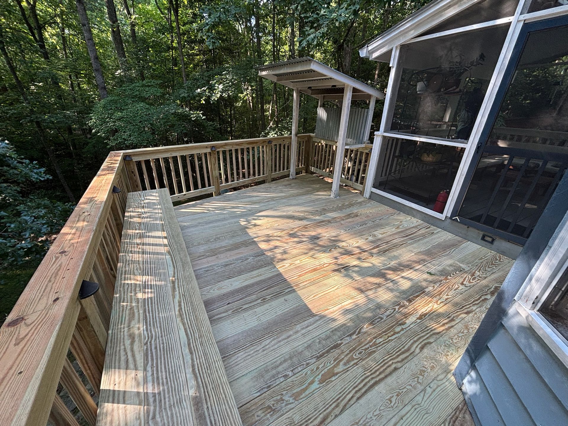 Wooden deck with railing and stairs beside a screened porch, surrounded by trees.
