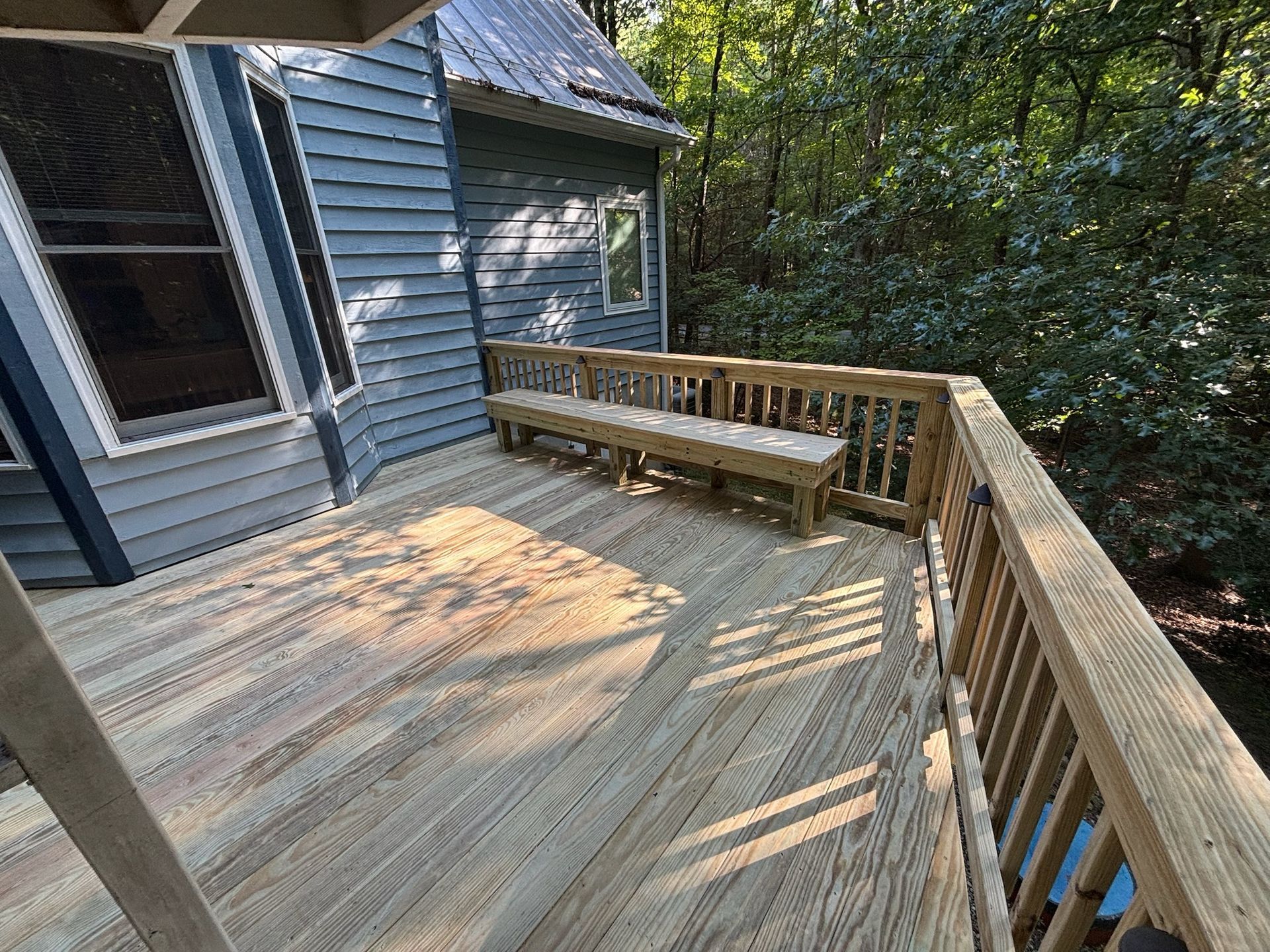 Wooden deck beside a blue house, with a bench and railing overlooking trees.