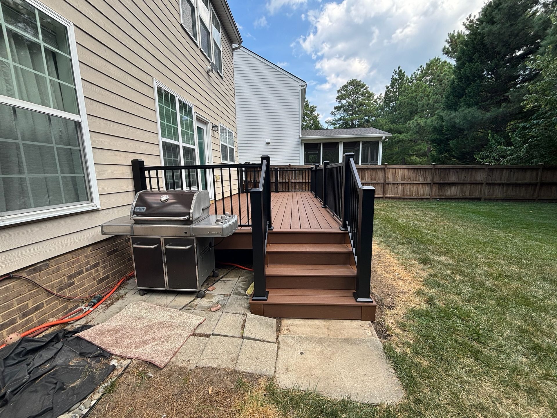 Backyard wooden deck steps beside a house, with black railings and a paved patio below