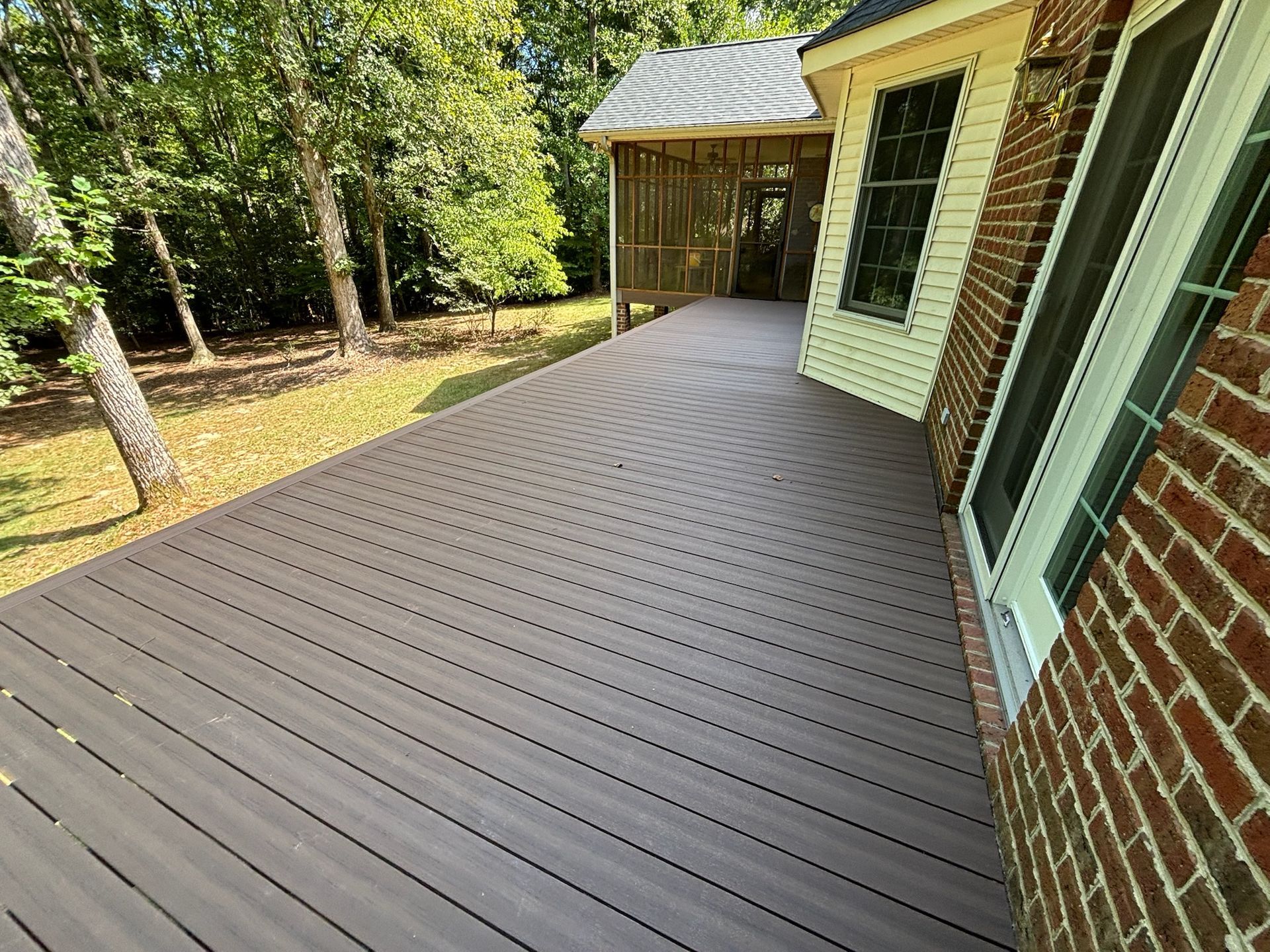 Wide brown composite deck beside a brick house, with a screened porch and trees in the backyard