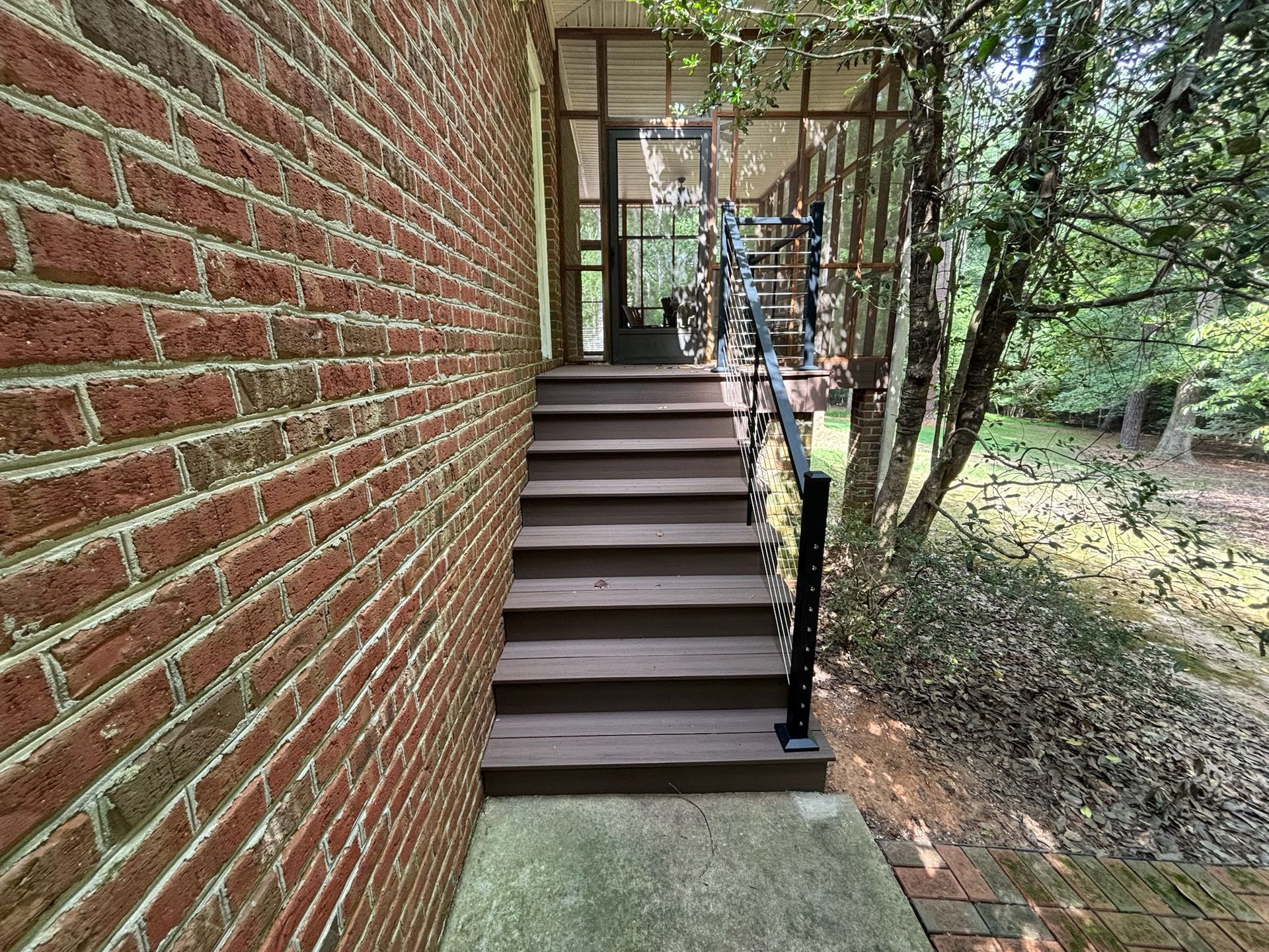 Brick house side entrance with stairs, black handrail, and surrounding trees