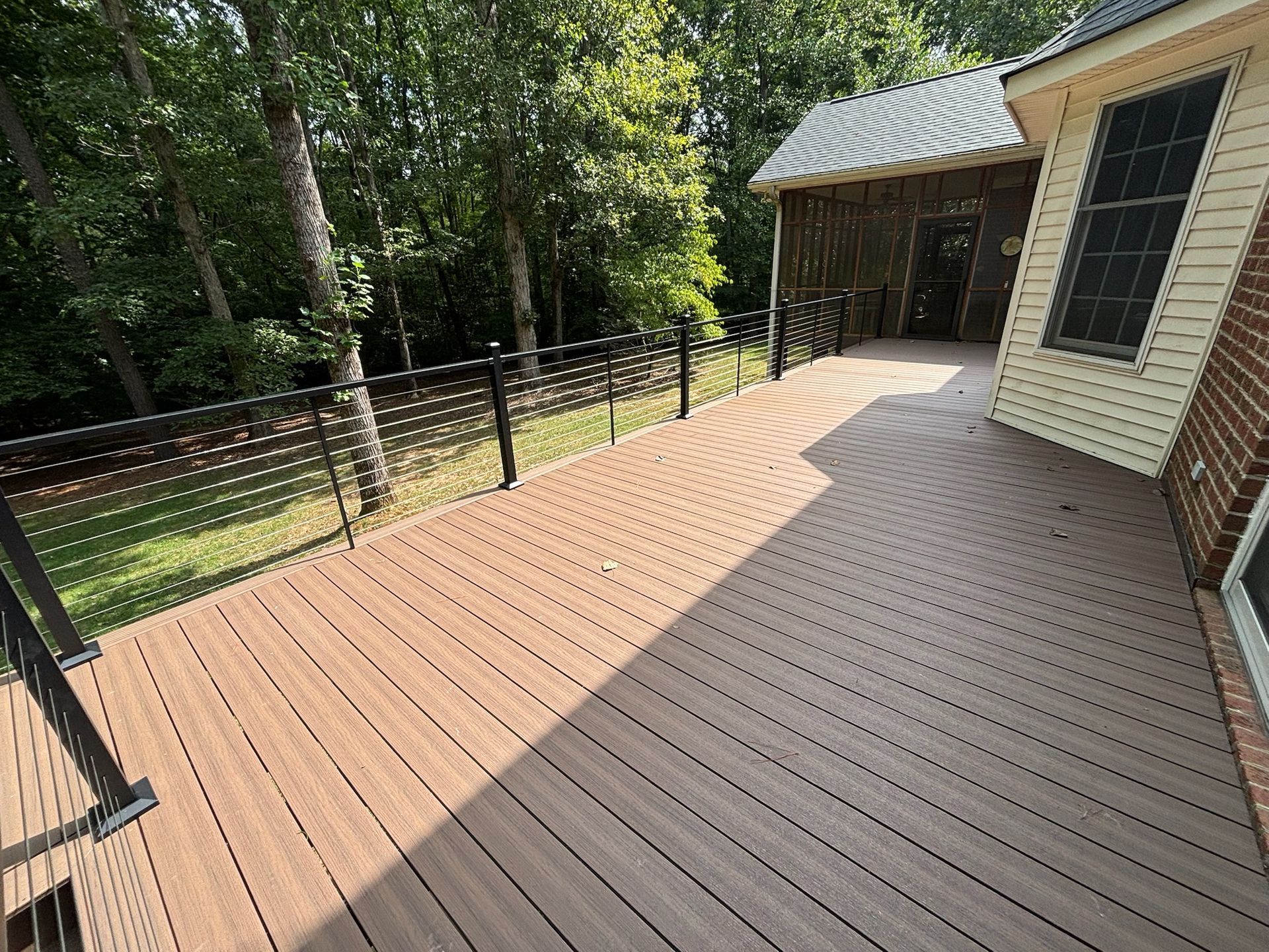 Wooden deck beside a brick house, with black railing and trees in the yard.