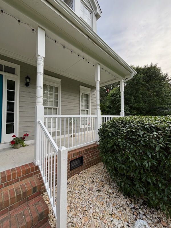 Front porch with white railing, brick steps, gravel path, and trimmed hedges beside a light-colored house