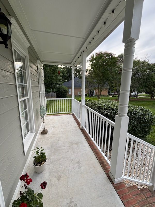Covered front porch with white railing, potted flowers, and view of a green lawn and street.