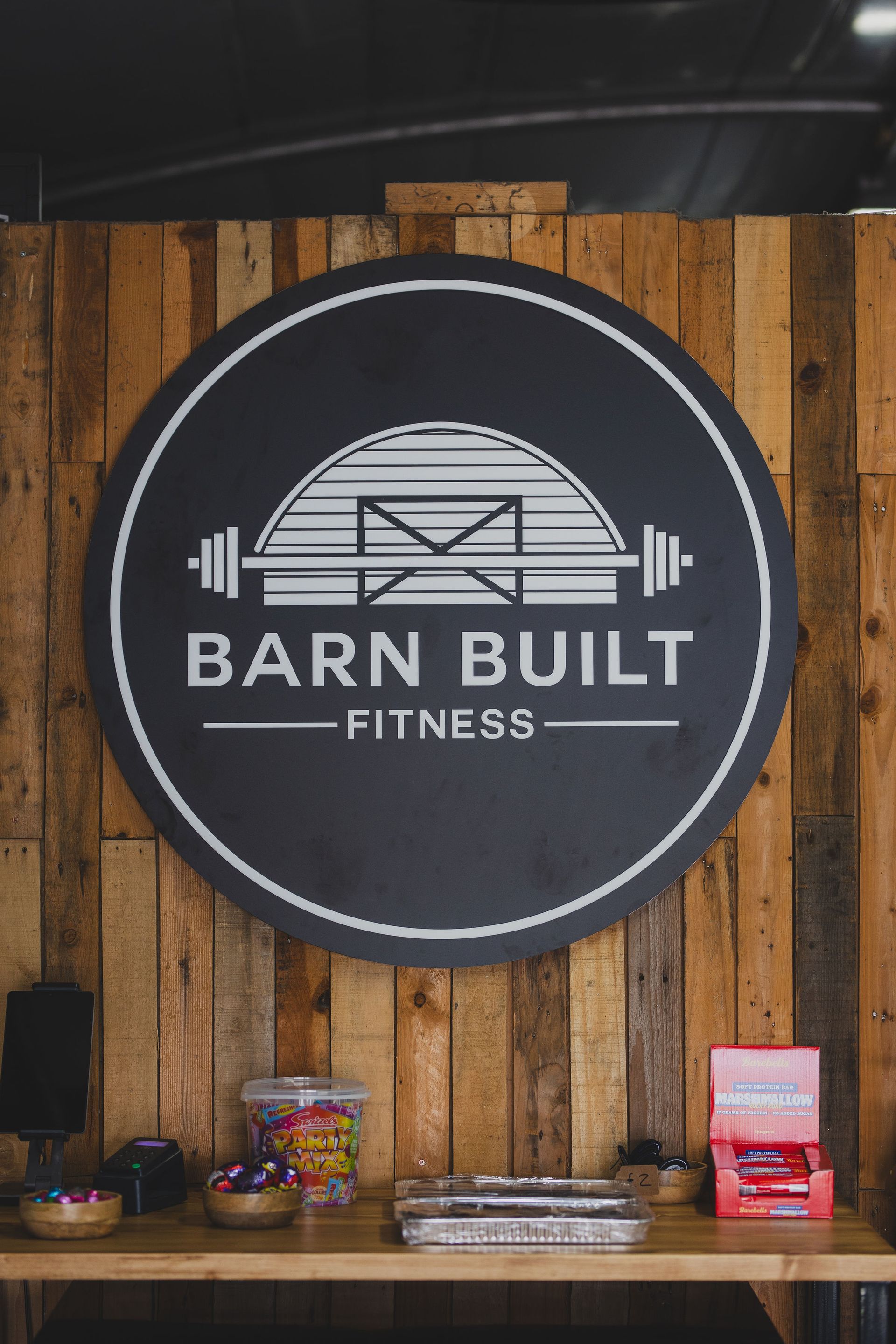 Barn Built Fitness sign on a wooden wall, with candy on a shelf below.