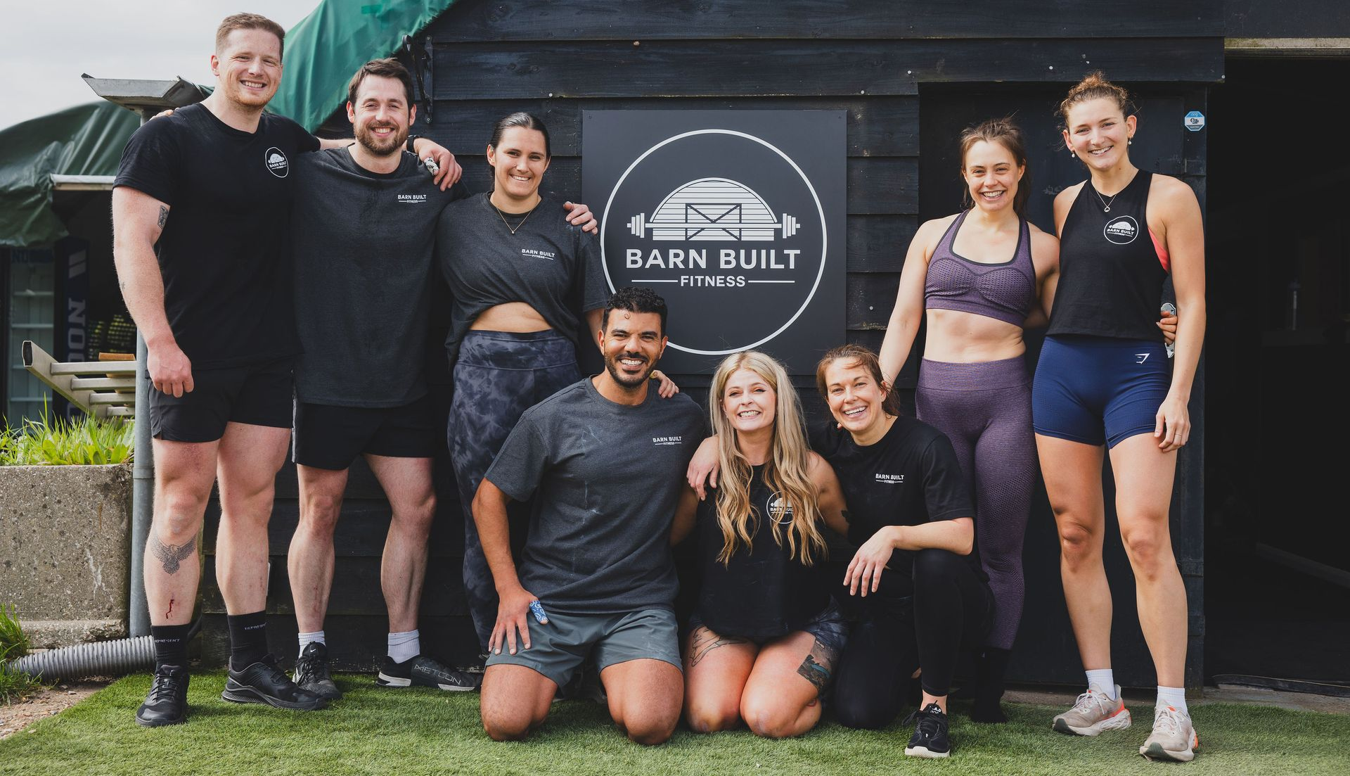 Group of people smiling in front Barn Built Fitness in Wokingham. Men in shorts, women in workout clothes. Outside.