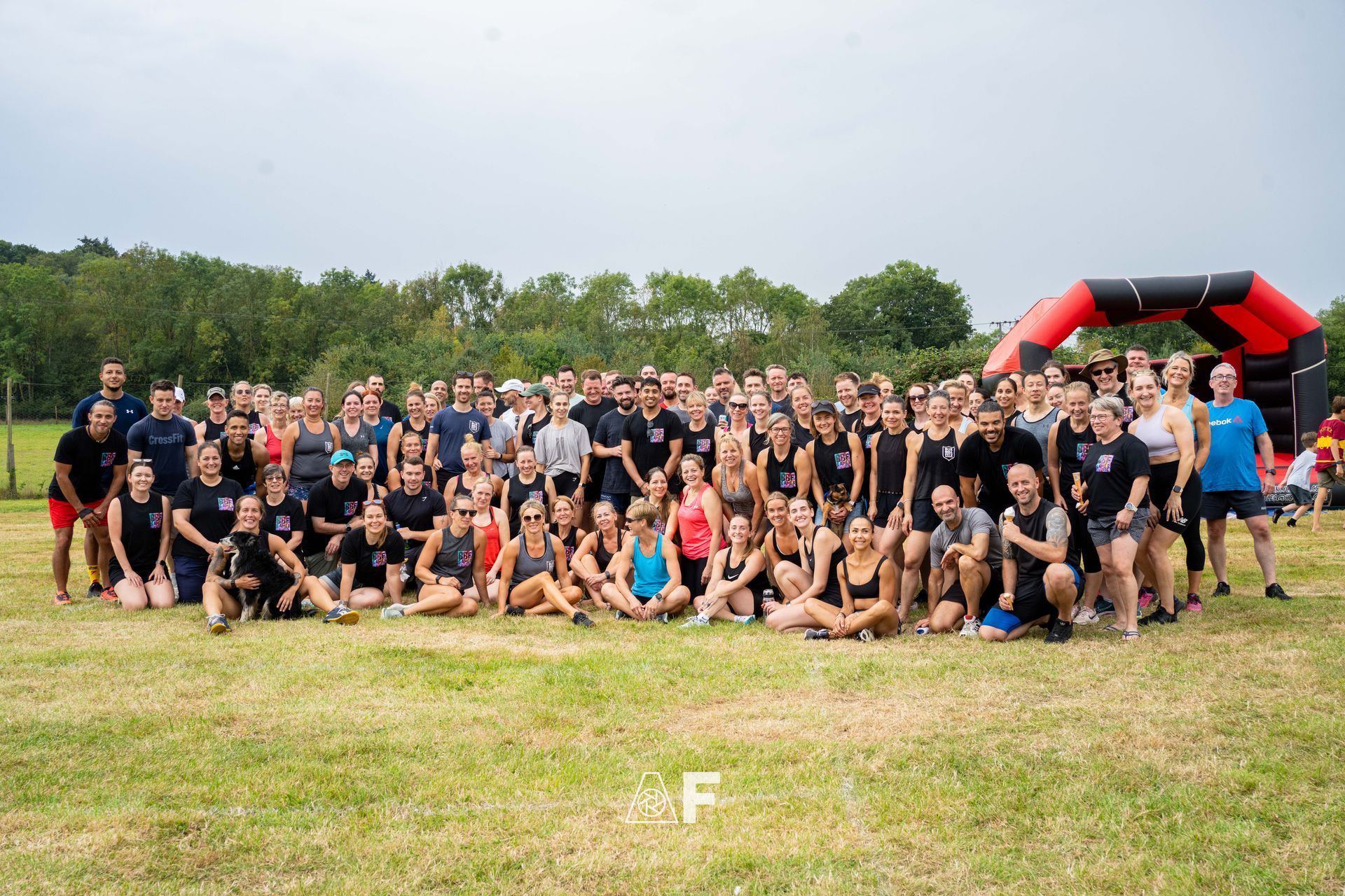Large group of people, diverse in age and build, posing on grass field. Red and black archway in background.