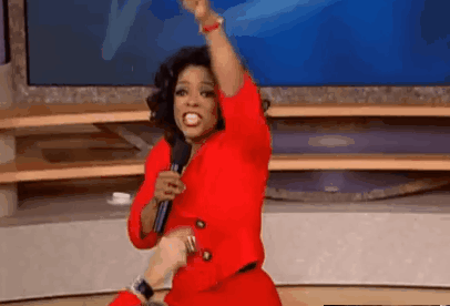 Oprah Winfrey raises an arm victoriously while holding a microphone; red blazer, blue backdrop.