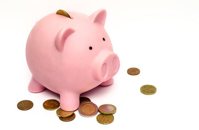 Pink piggy bank with a coin in the slot, surrounded by loose change on a white surface.
