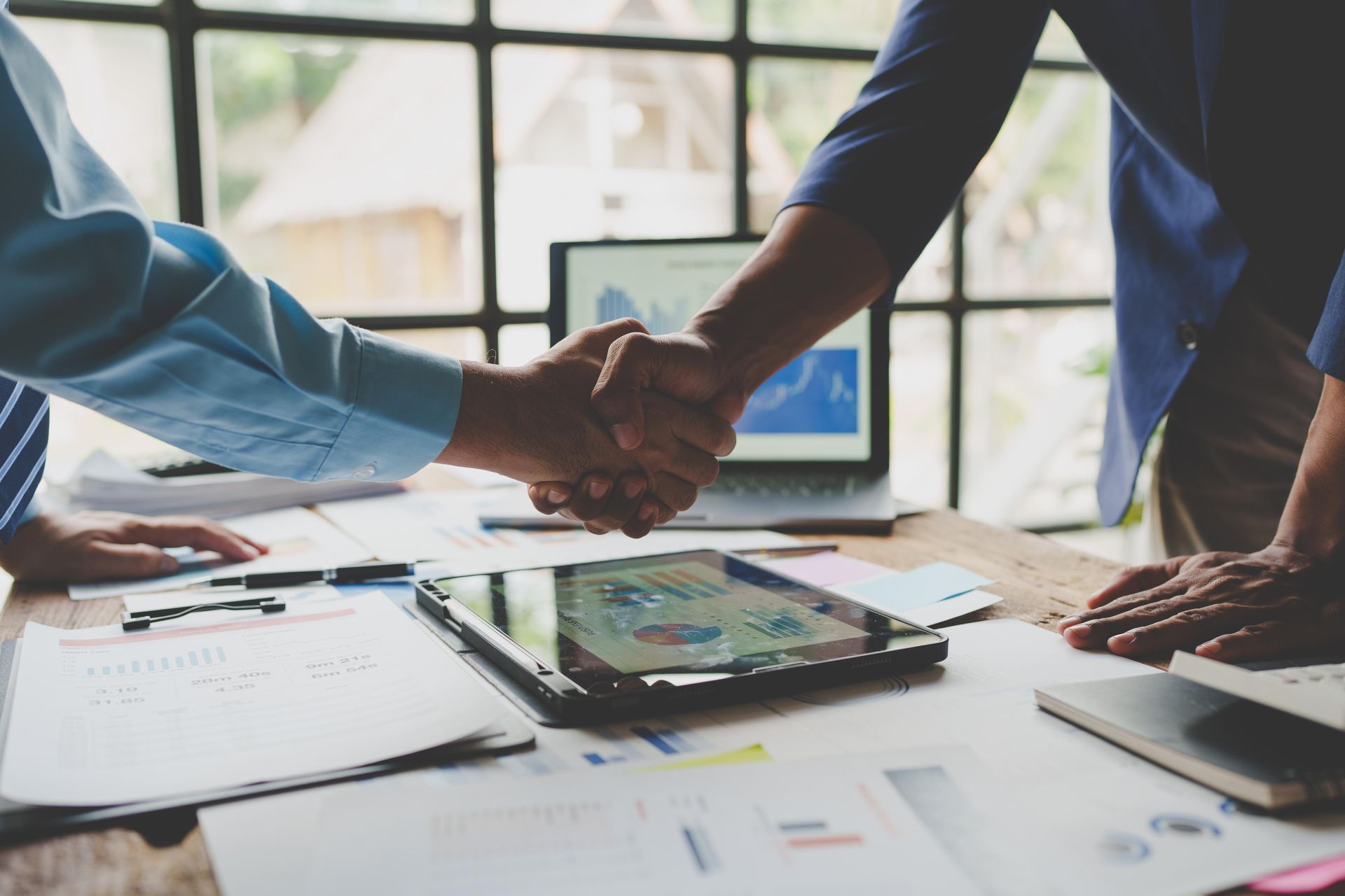 Two people shaking hands over a desk with documents, tablet, and laptop displaying charts.