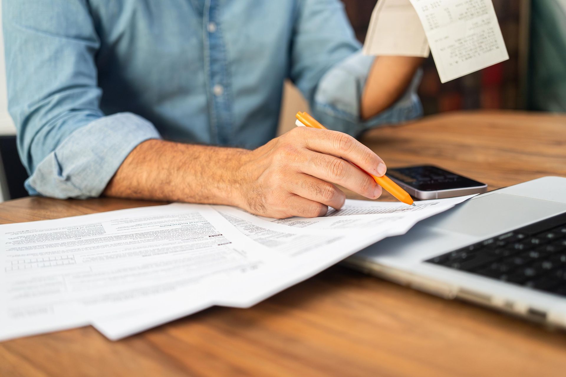 Person reviewing documents, holding a receipt, using a pen, and working on a laptop.
