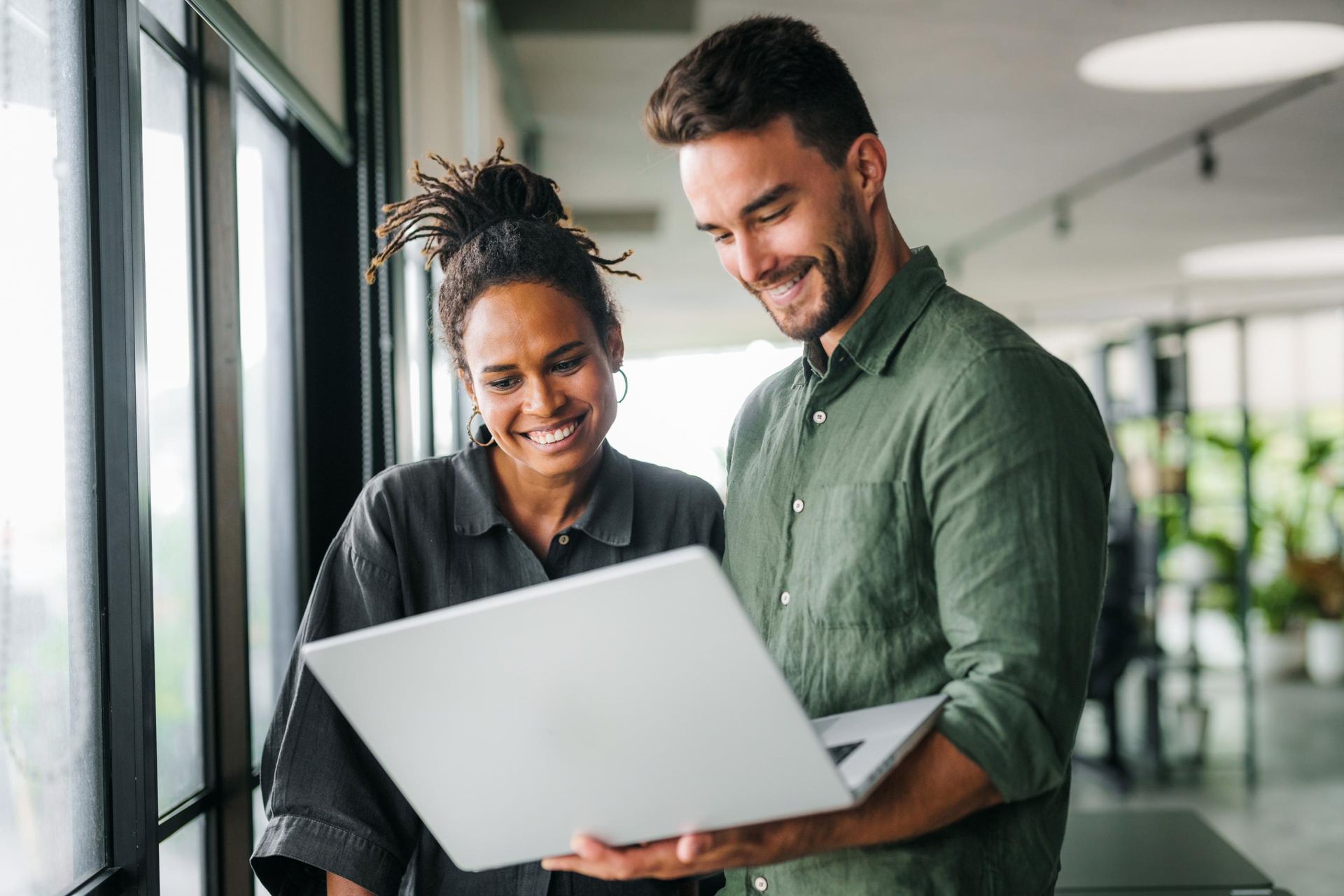 Two people looking at laptop, smiling near a window. 