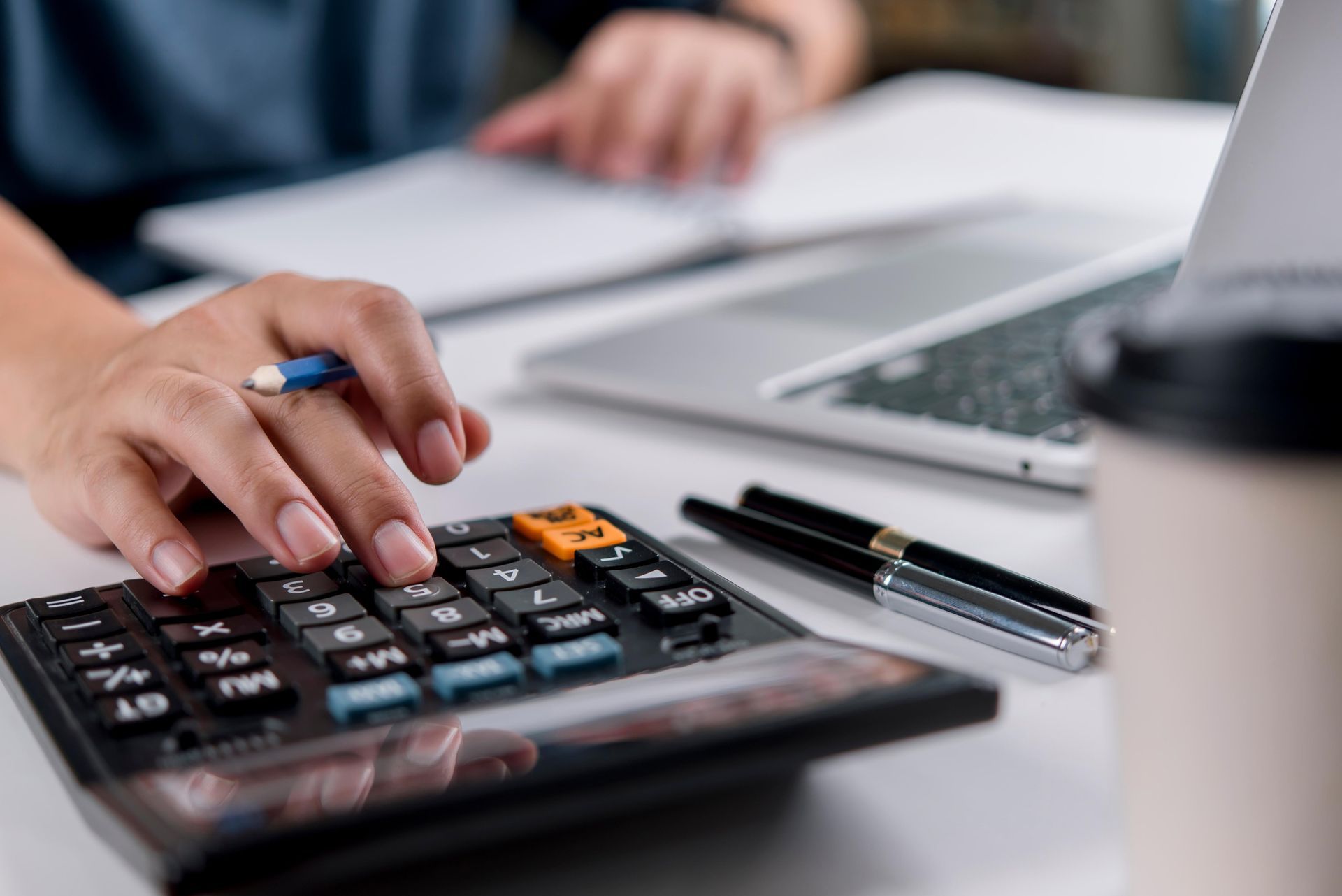 Person's hand using a calculator, notebook and laptop in background.