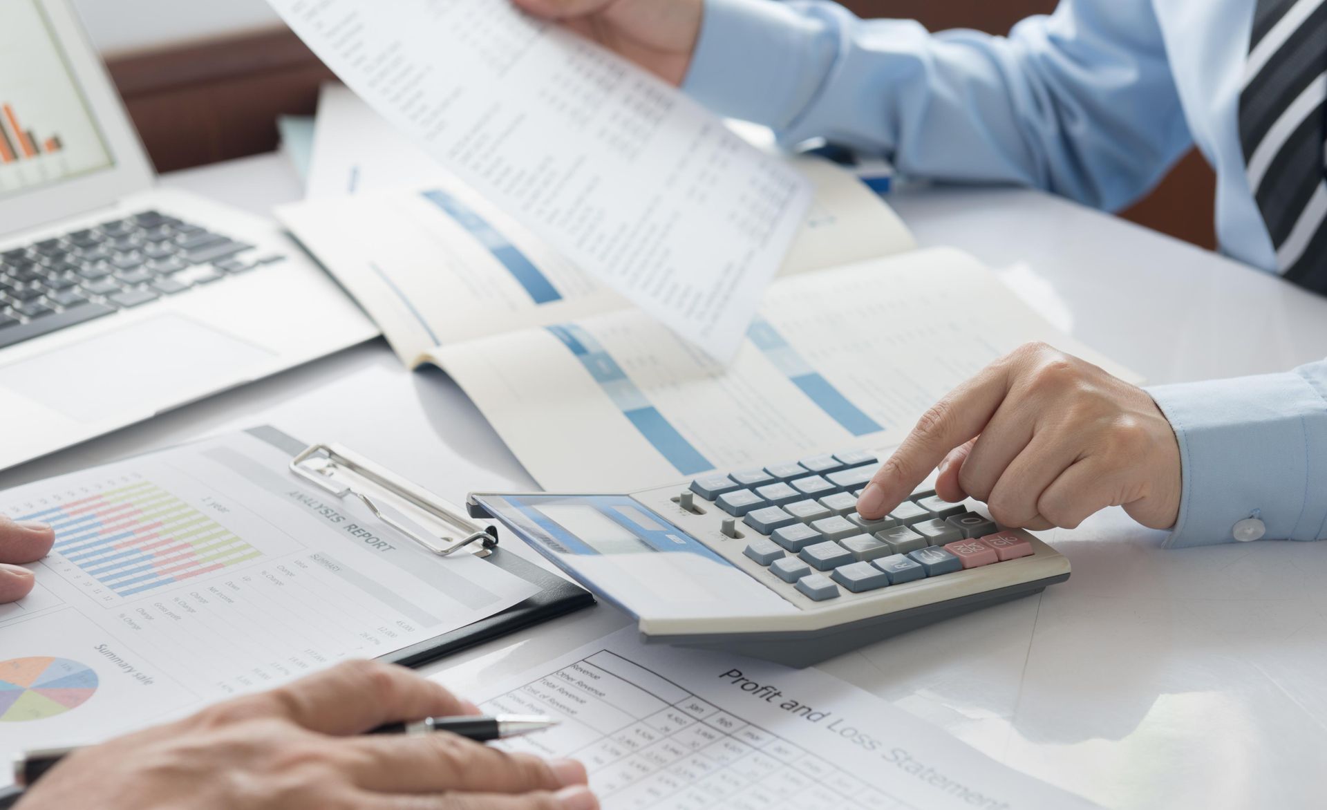 Two people review financial documents, one using a calculator, papers and a laptop on a white table.