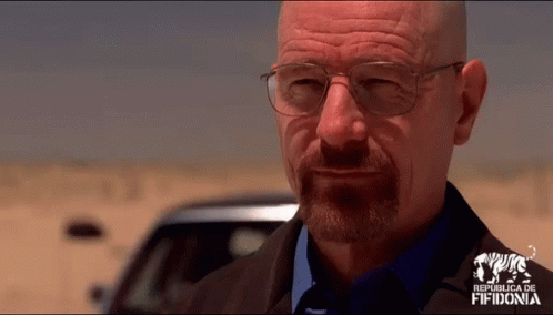 Bald man in glasses and goatee smirks, standing in a desert, with a car in the background.