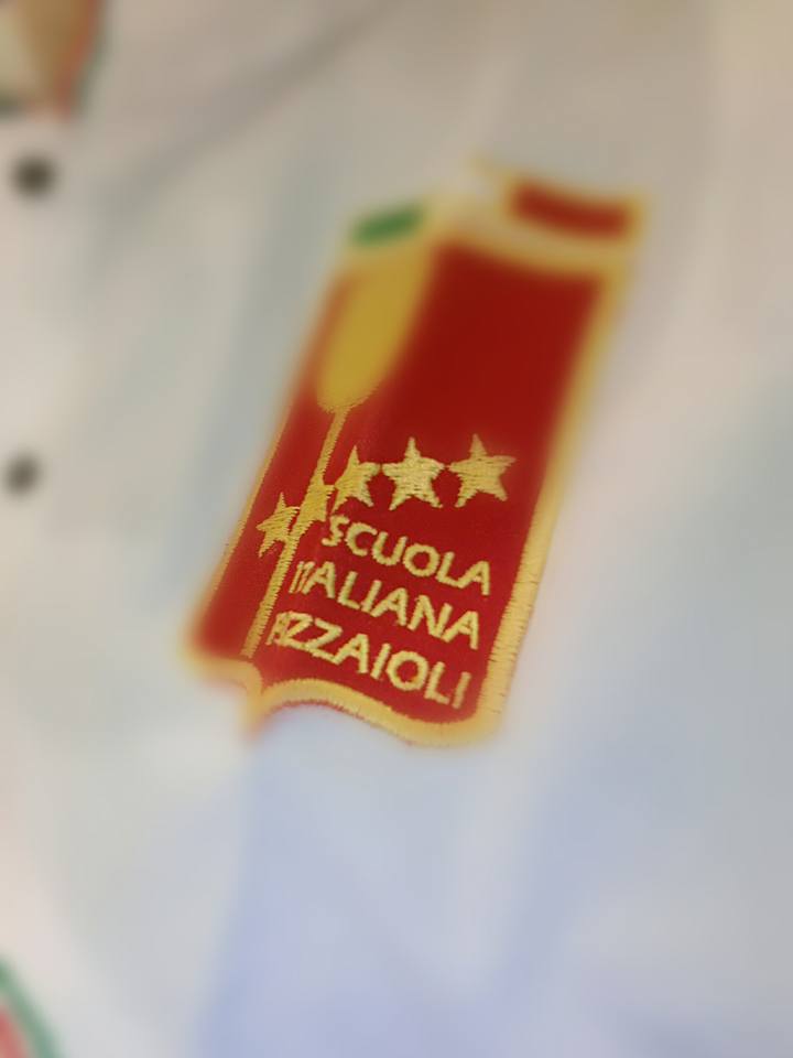 Stemma della Scuola Italiana Pizzaioli ricamato in rosso e oro su una maglietta bianca.
