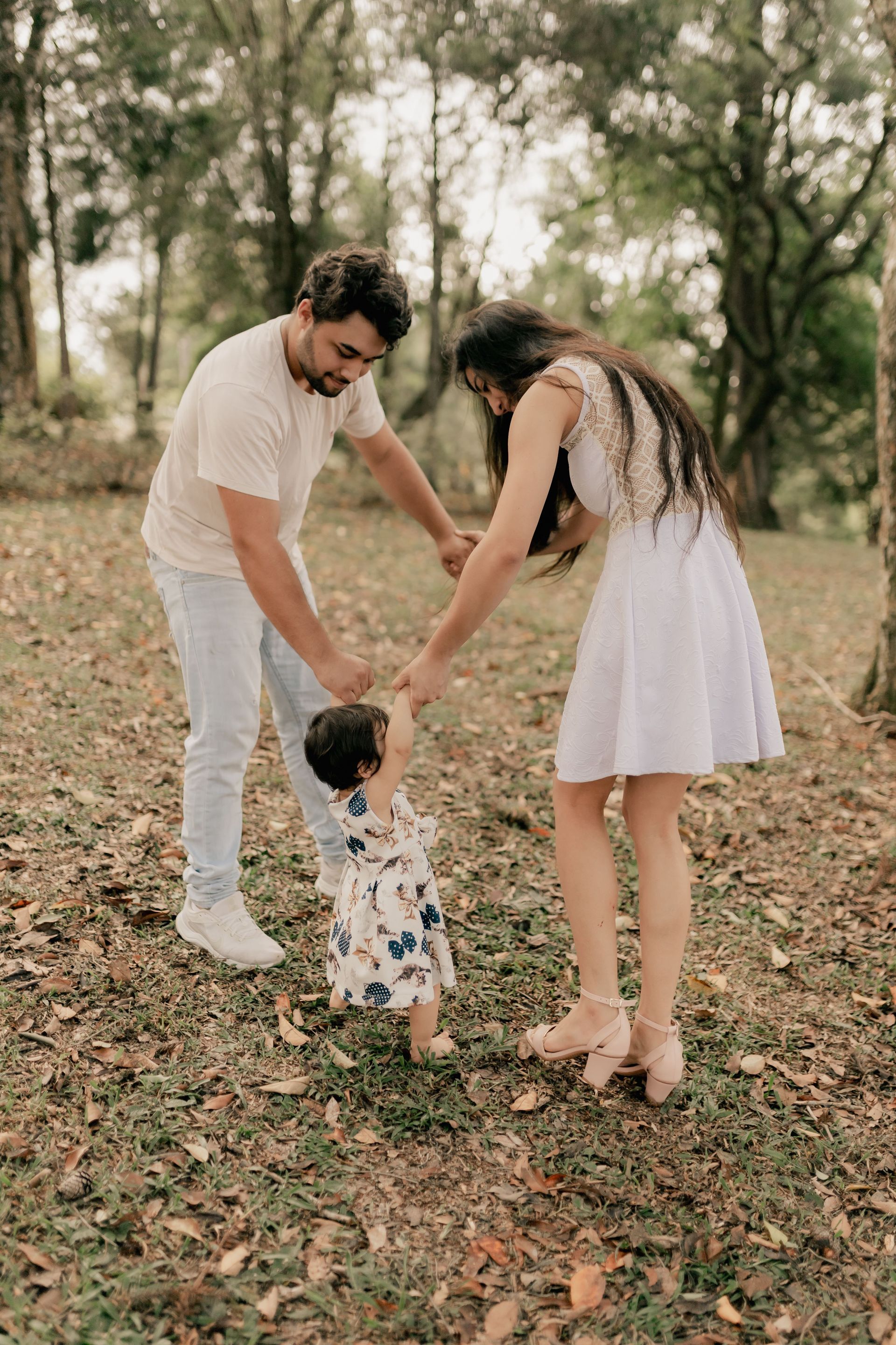 Family holding a toddler’s hands in a leafy park, with soft sunlight and trees in the background.