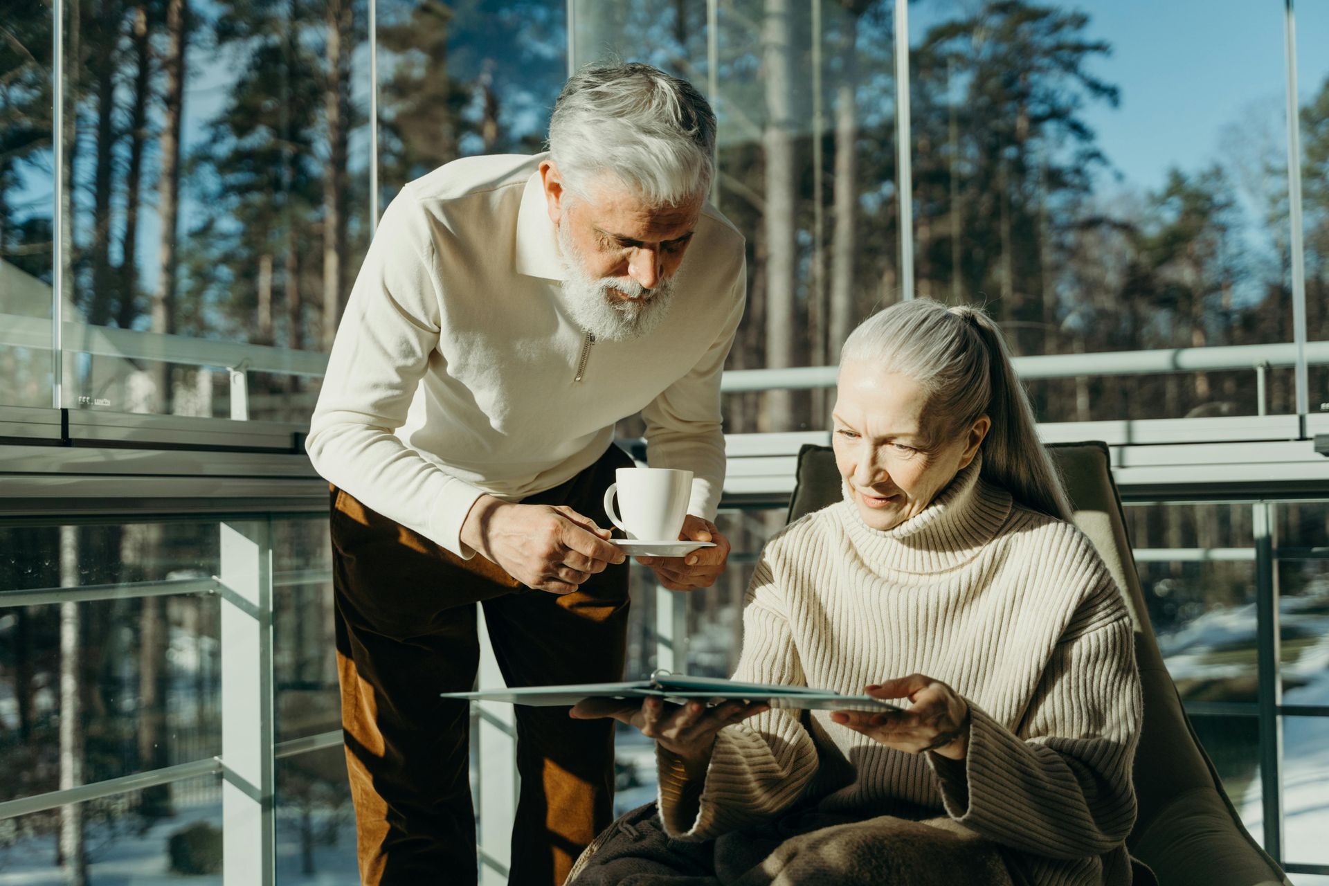 Two people sharing coffee on a sunlit outdoor patio, one seated with a blanket and looking at a tablet.