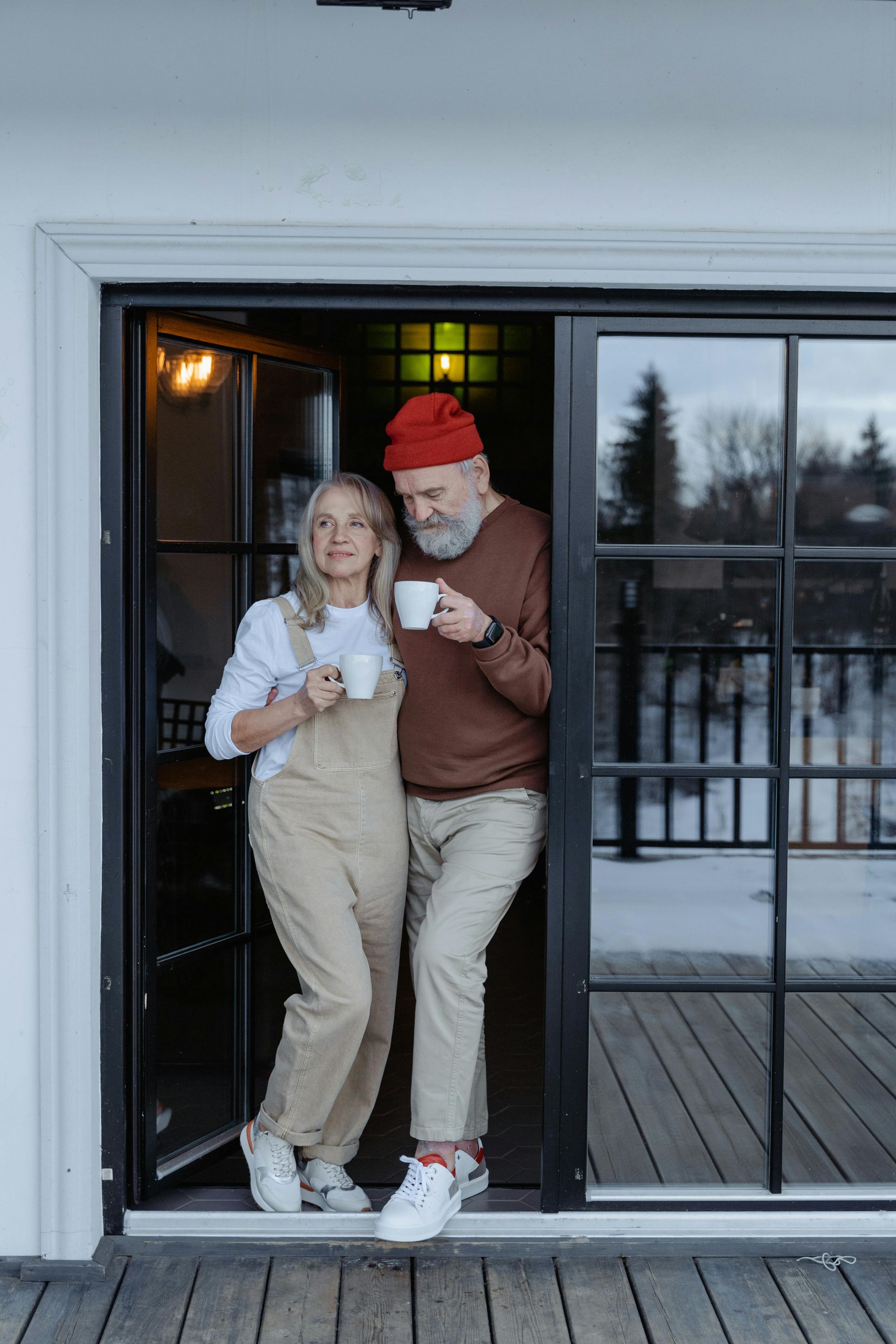 Two people stand in a doorway holding drinks, wearing winter clothes, with snow outside.