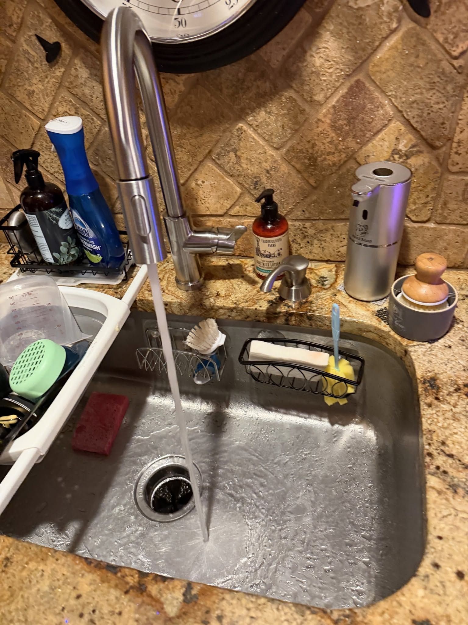Kitchen sink with running faucet, dish soap, sponge, and cleaning supplies on the counter.