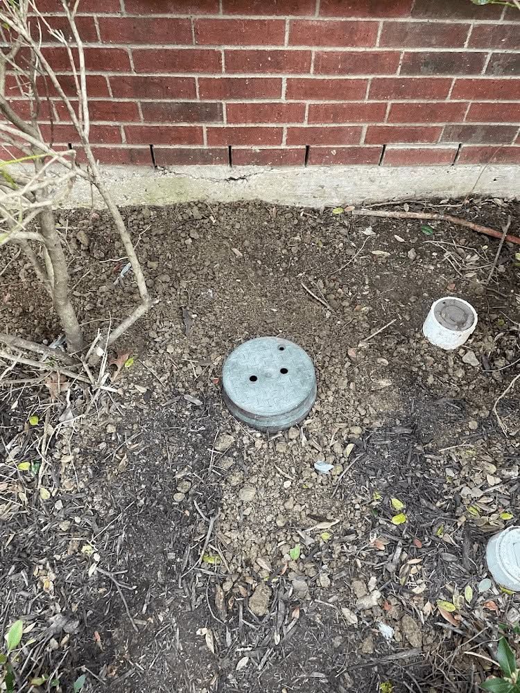 Ground-level garden utility cap beside a brick wall, with soil and small plants around it.