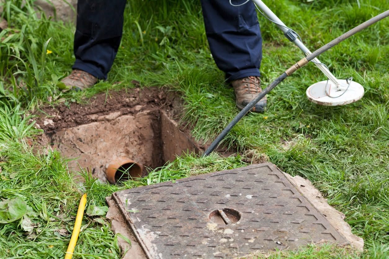 Workers inspecting an open manhole in a grassy area, with a metal cover removed nearby.