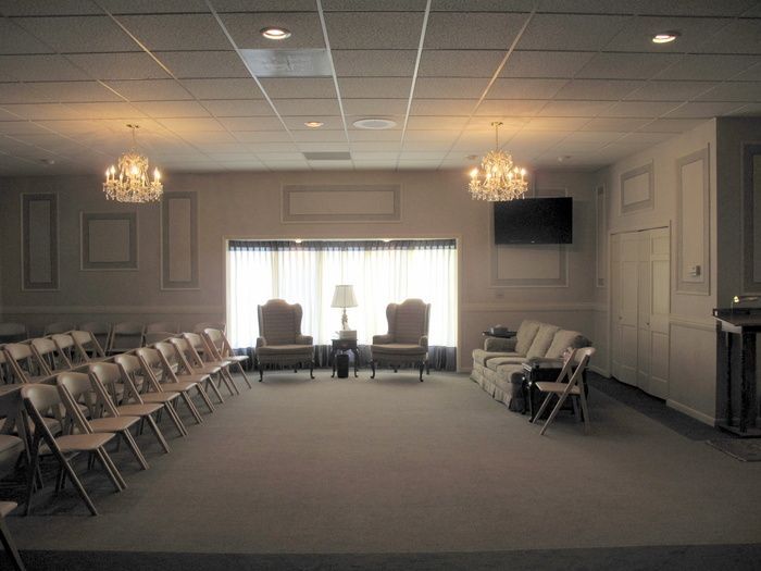 Empty meeting room with chairs lining the walls, chandeliers, and bright windows at the far end.
