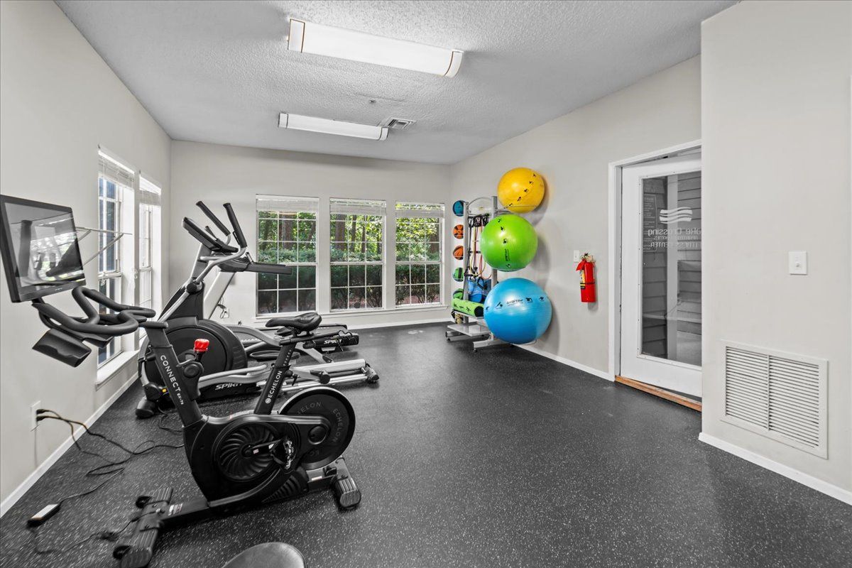 A brightly lit apartment fitness center with stationary bikes, exercise balls in a rack, and a glass-paneled door.