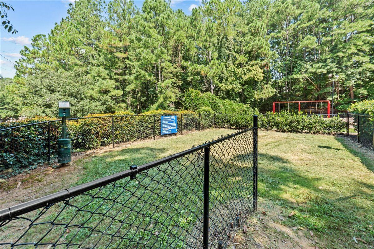 A fenced-in dog park with a grassy lawn, a waste station, a blue sign, and a swing set in the background.