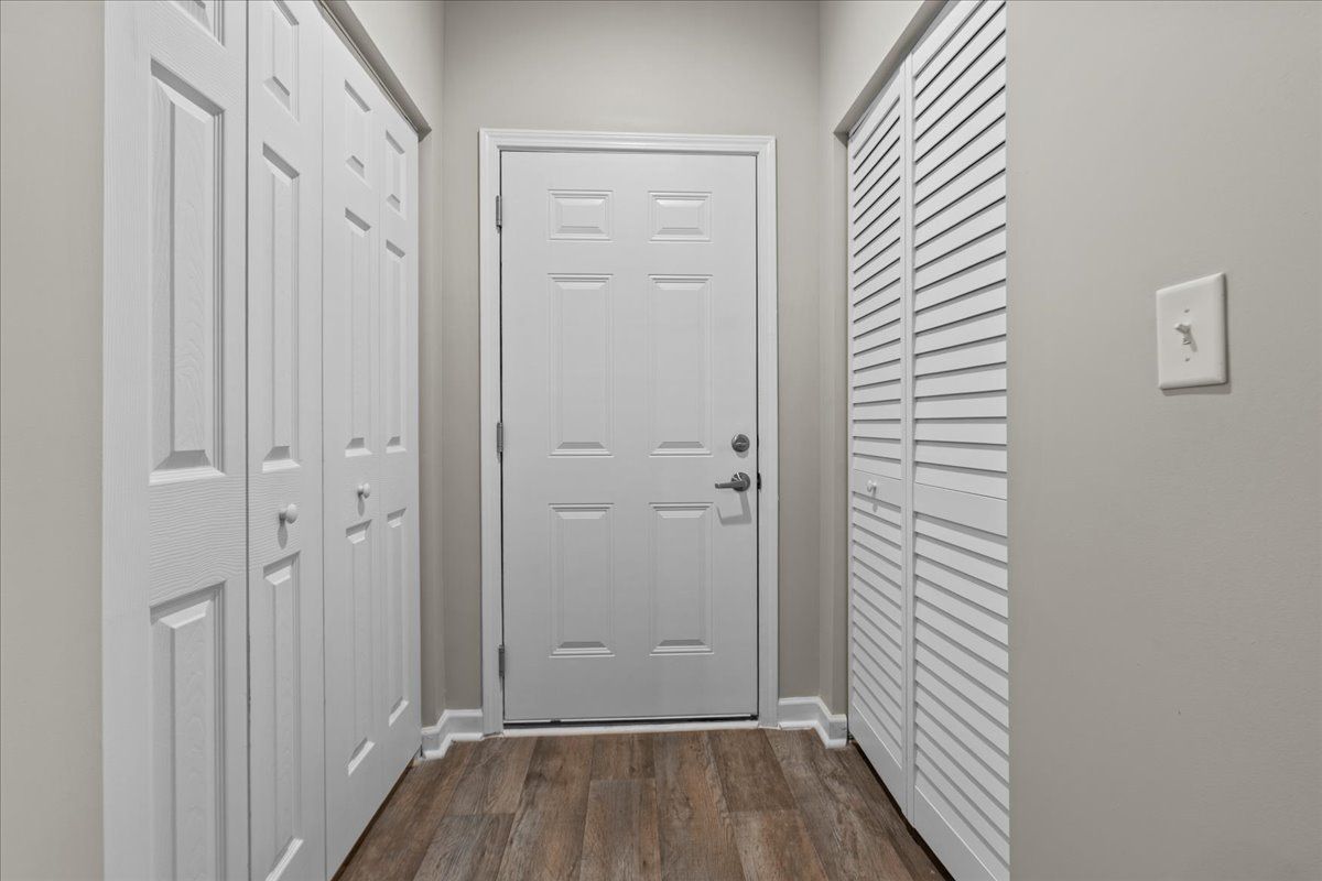 A narrow interior entryway with a white door, louvered closet doors on the right, solid doors on the left, and wood floors.