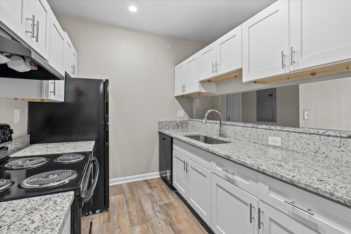 A galley-style kitchen with white cabinets, speckled granite countertops, a black stove, and light wood-look flooring.