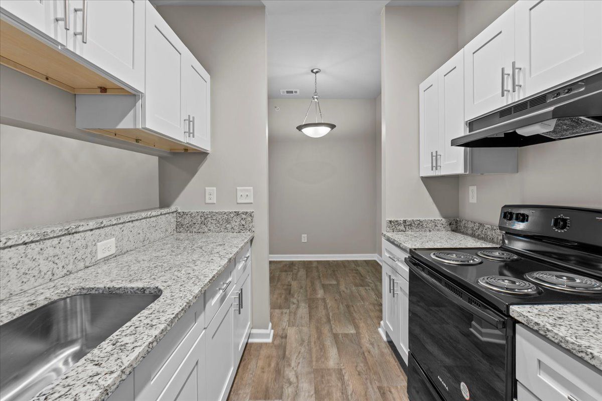 A bright galley kitchen with white cabinets, granite countertops, stainless steel sink, black range, and wood floors.