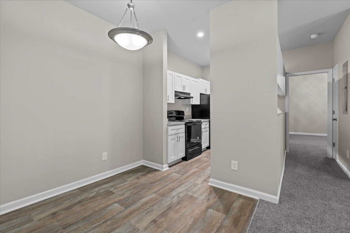 A dining area with wood-look flooring leads to a white kitchen with black appliances, adjacent to a carpeted hallway.