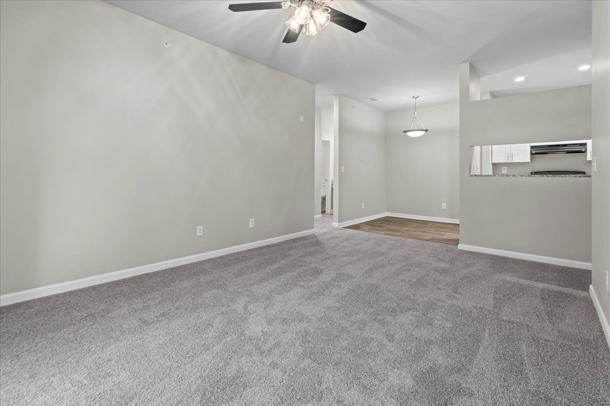 A spacious, empty living room with gray carpet, neutral walls, a ceiling fan, and an open view into a kitchen area.