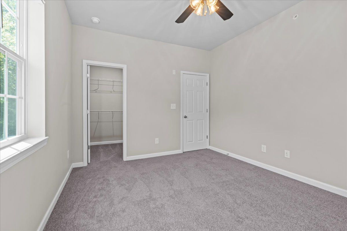Empty bedroom with grey carpet, beige walls, a window on the left, a walk-in closet, and a white bedroom door.