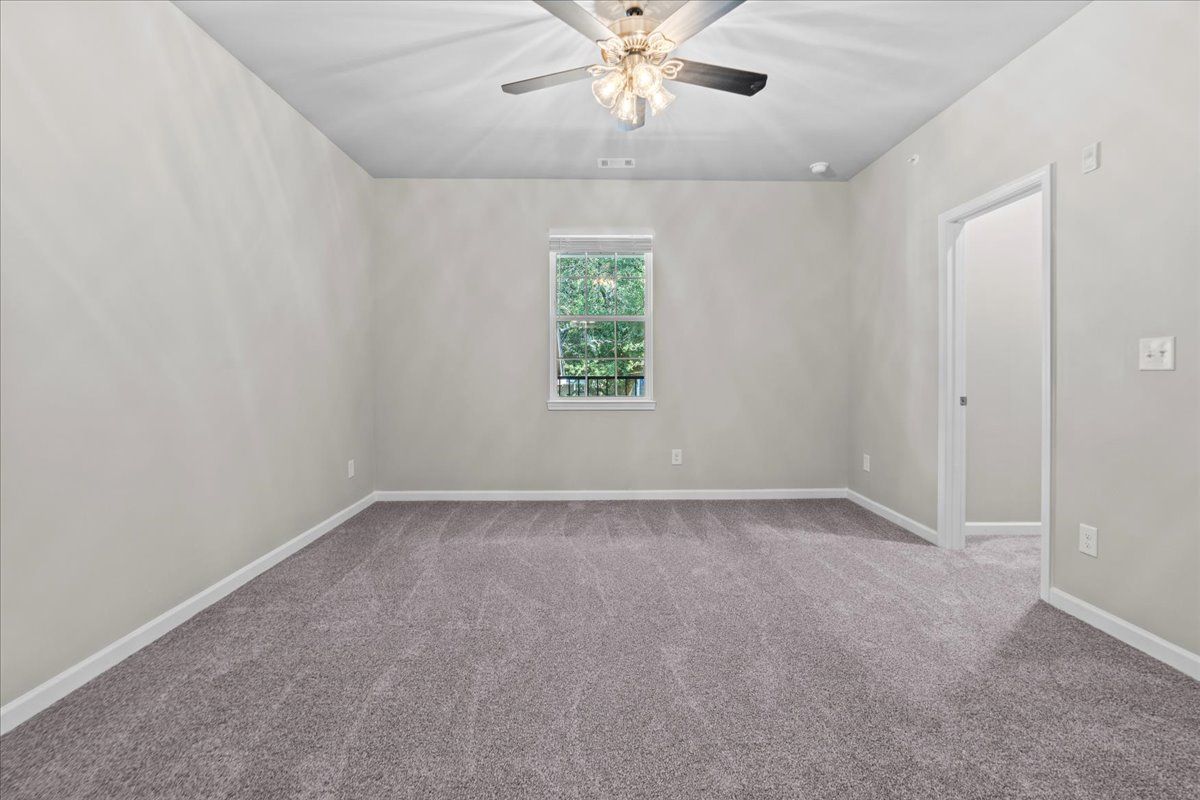 A bright, empty bedroom with gray carpet, light gray walls, a ceiling fan, and a doorway leading to another room.