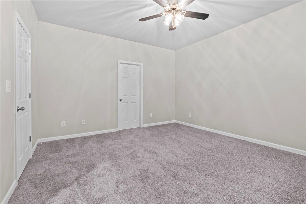A sparsely furnished bedroom with light gray walls, carpeted floors, two white interior doors, and a ceiling fan.