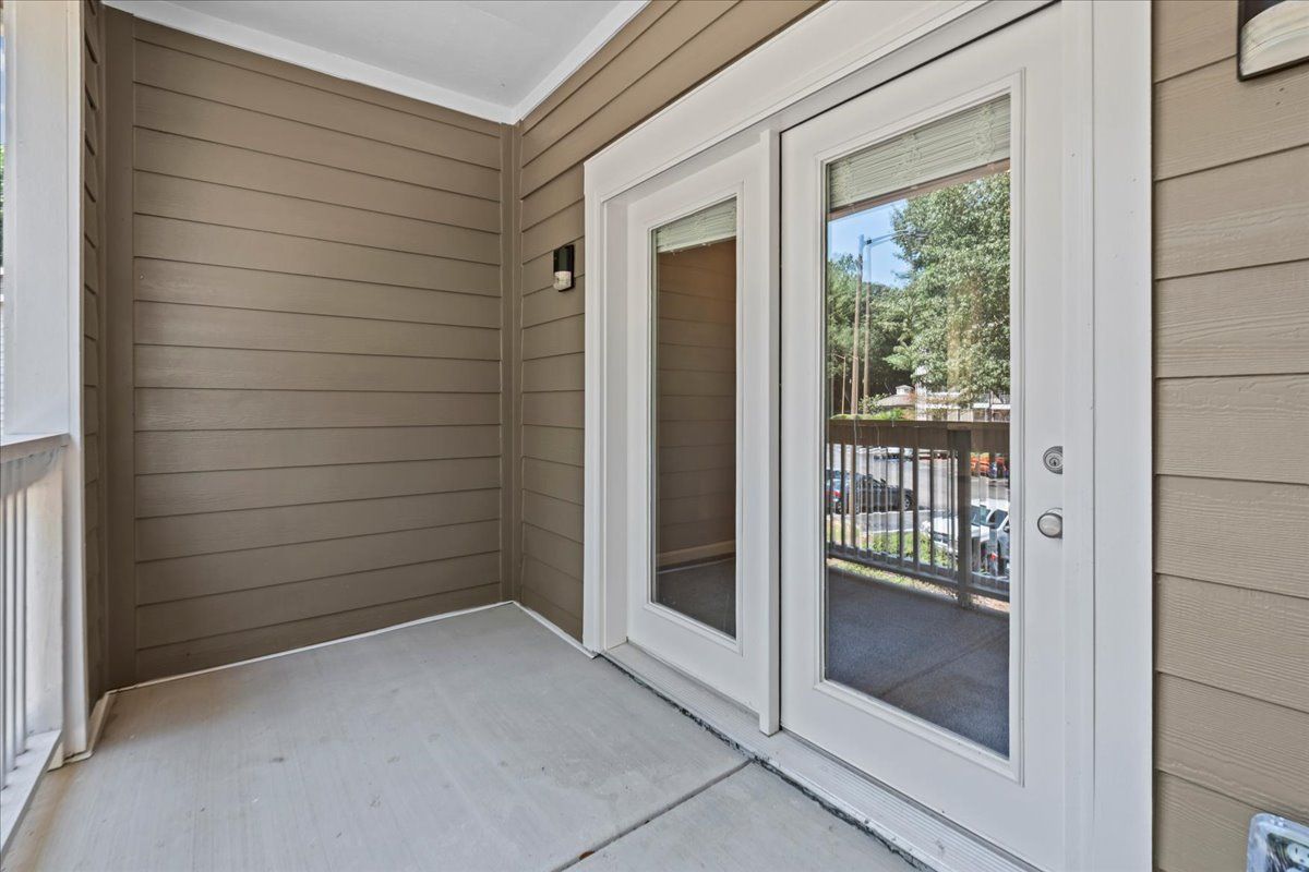 A small covered patio with light concrete flooring and brown horizontal siding, featuring a white double-glass door.