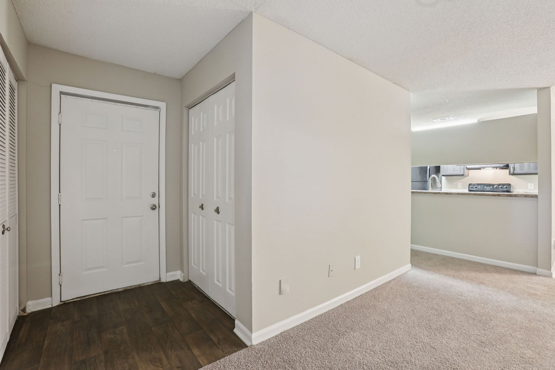 An entryway with a white door and beige walls, transitioning from dark wood flooring to carpet, with a kitchen view.