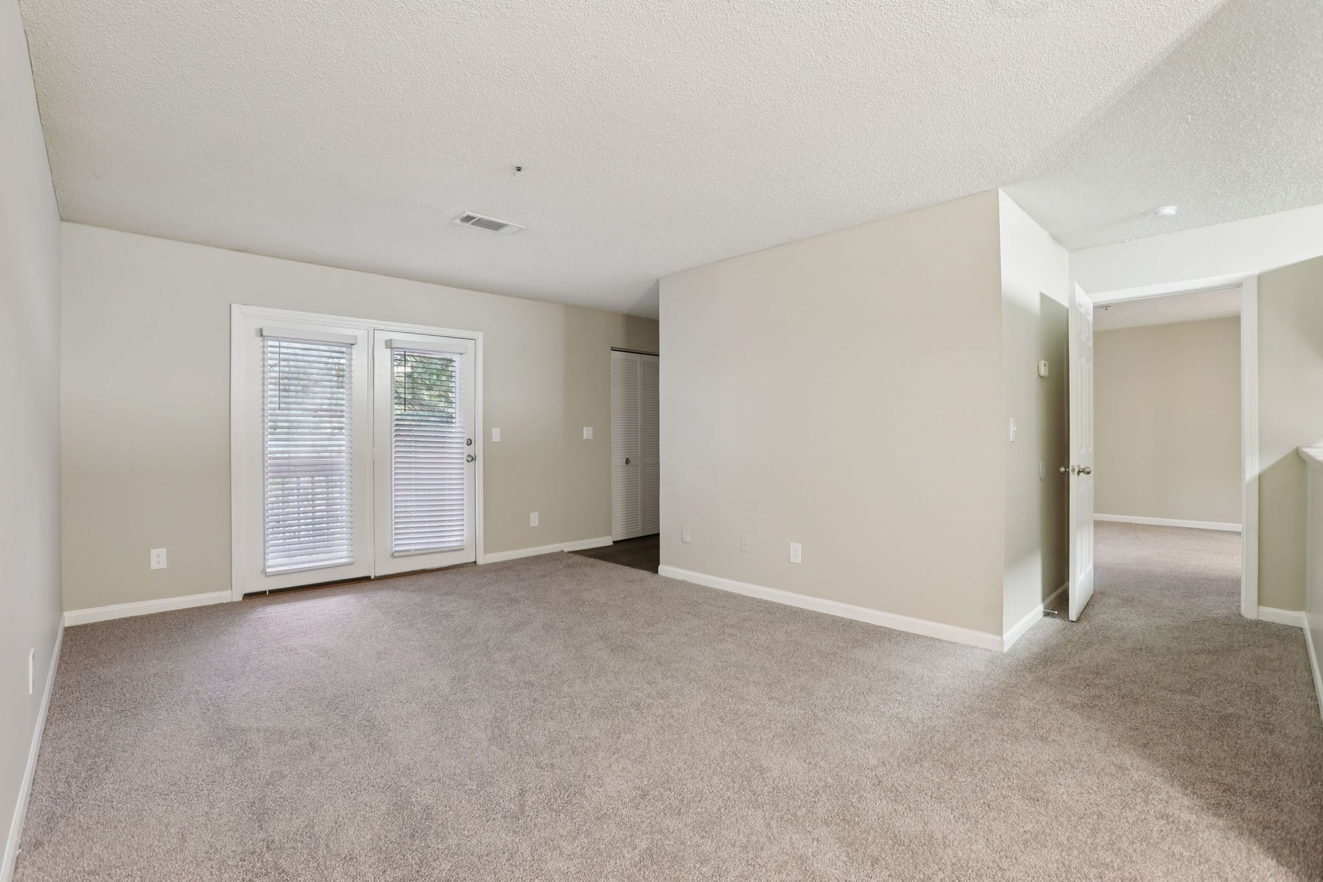 An empty room with neutral walls, tan carpeting, and French doors leading outdoors.