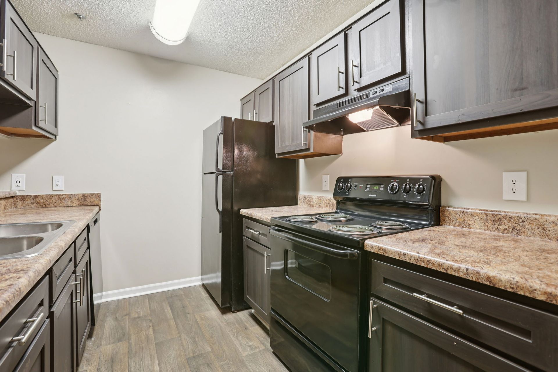 A narrow kitchen featuring dark wood cabinets, black appliances, tan countertops, and light gray wood-look flooring.
