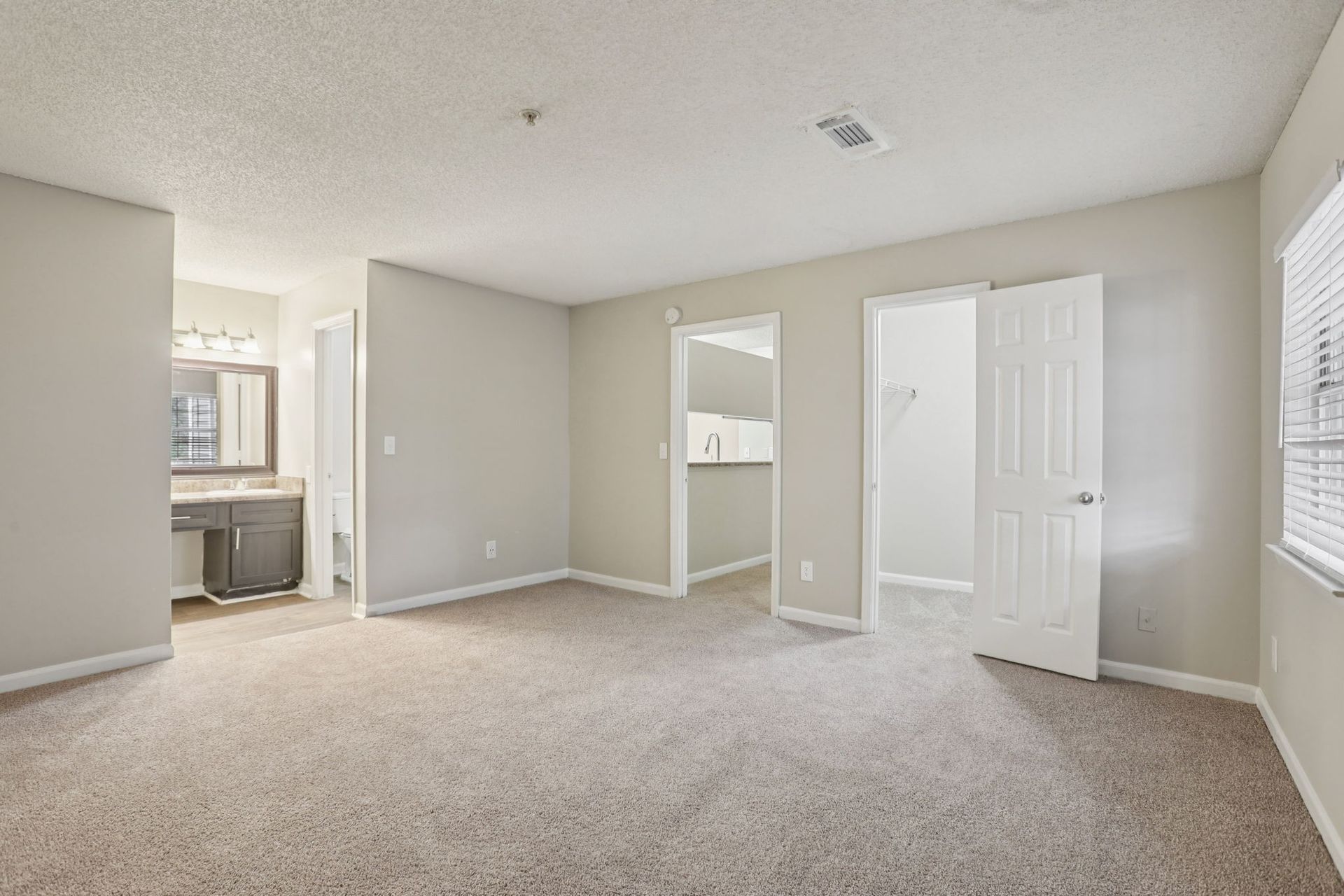 A spacious, empty bedroom with neutral walls, beige carpet, a vanity area, and two doorways leading to a bathroom and closet.