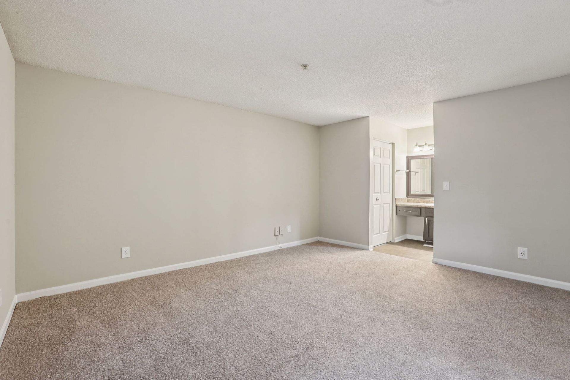 An empty bedroom with light gray walls, carpeted flooring, a popcorn ceiling, and an open doorway to a bathroom.