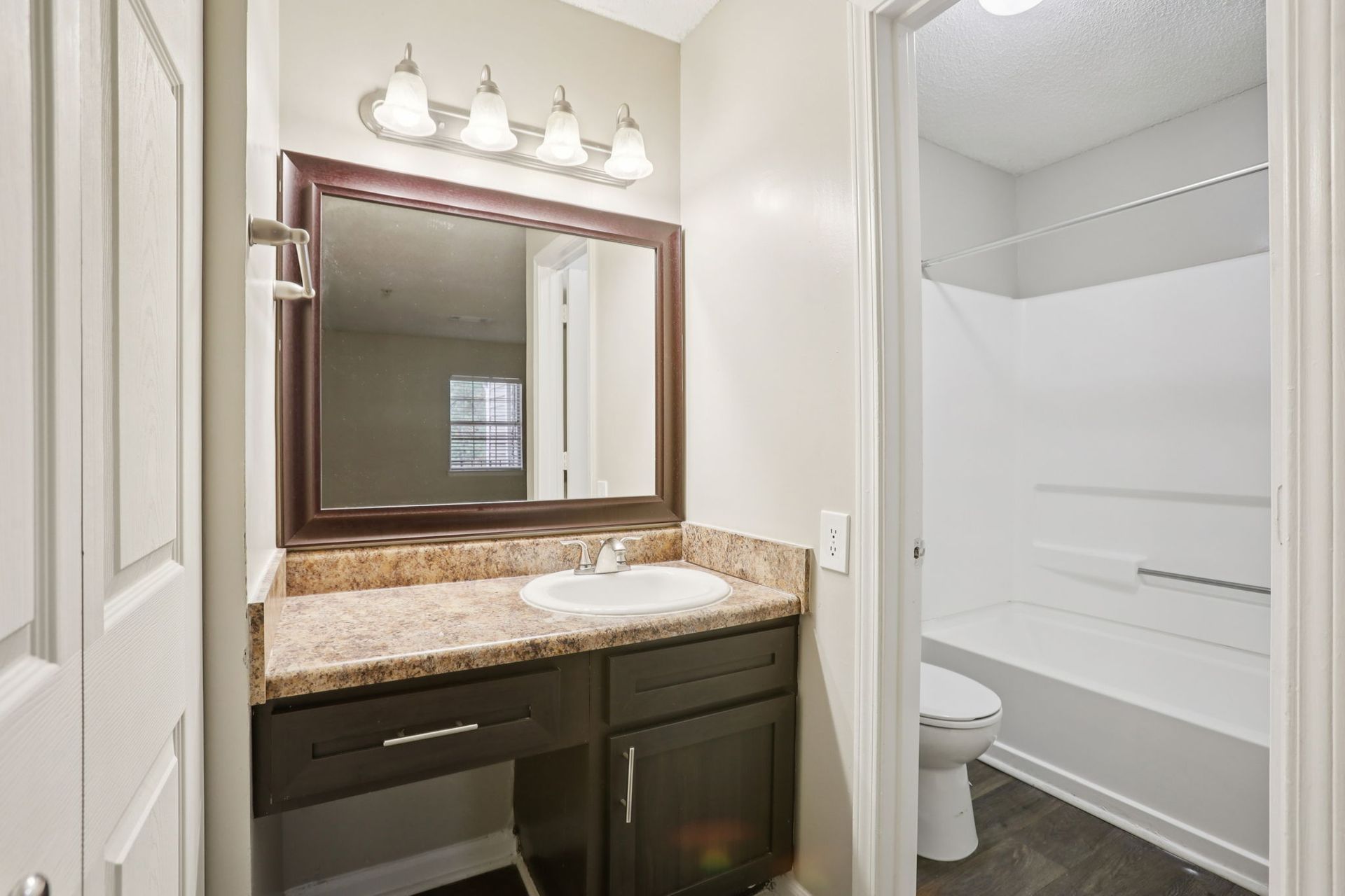 A small, modern bathroom with dark wood cabinets, a tan granite countertop, white sink, mirror, and a walk-in shower.