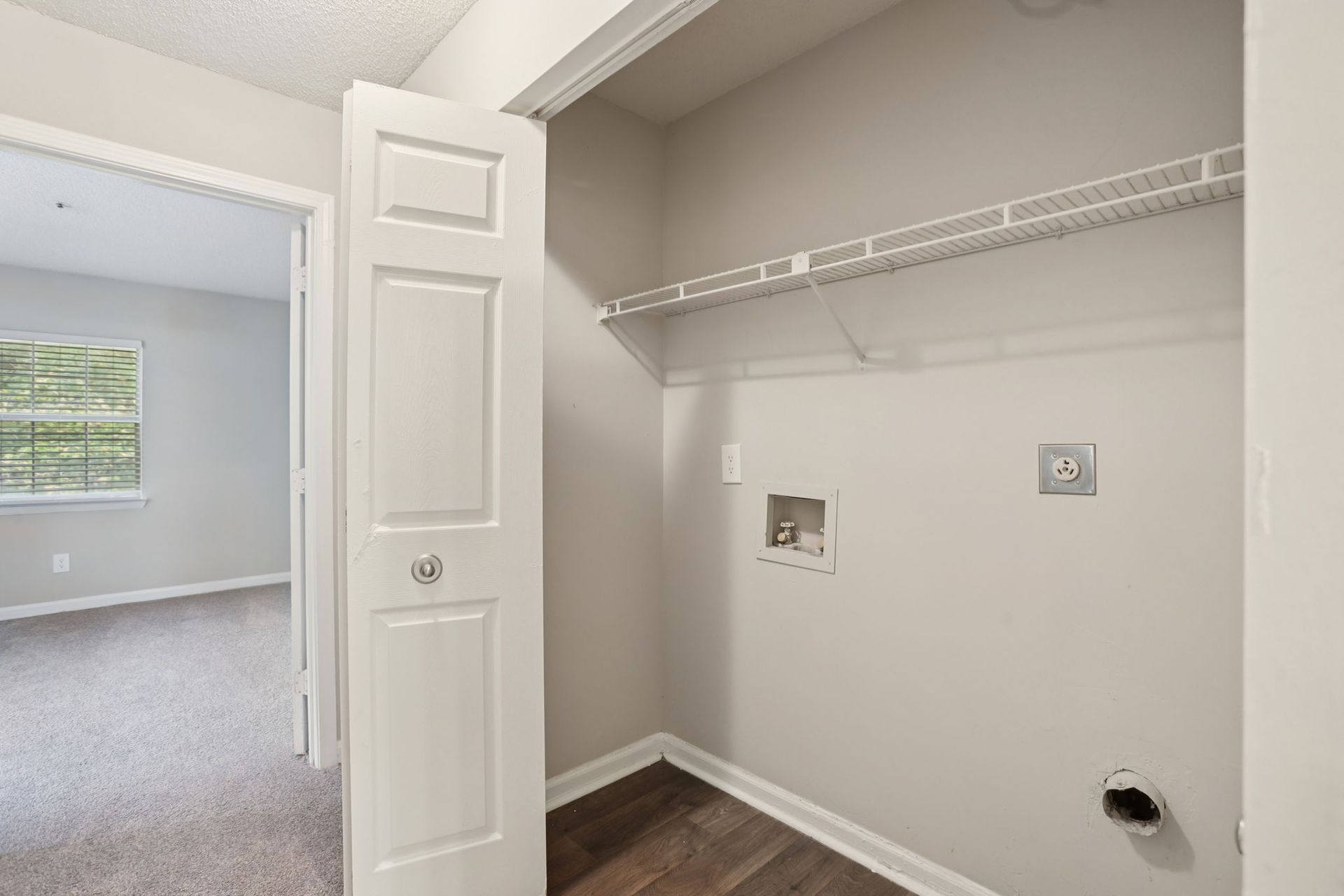 A neutral-toned laundry closet with a wire shelf, plumbing hookups, and a vent, adjacent to a carpeted room with a window.