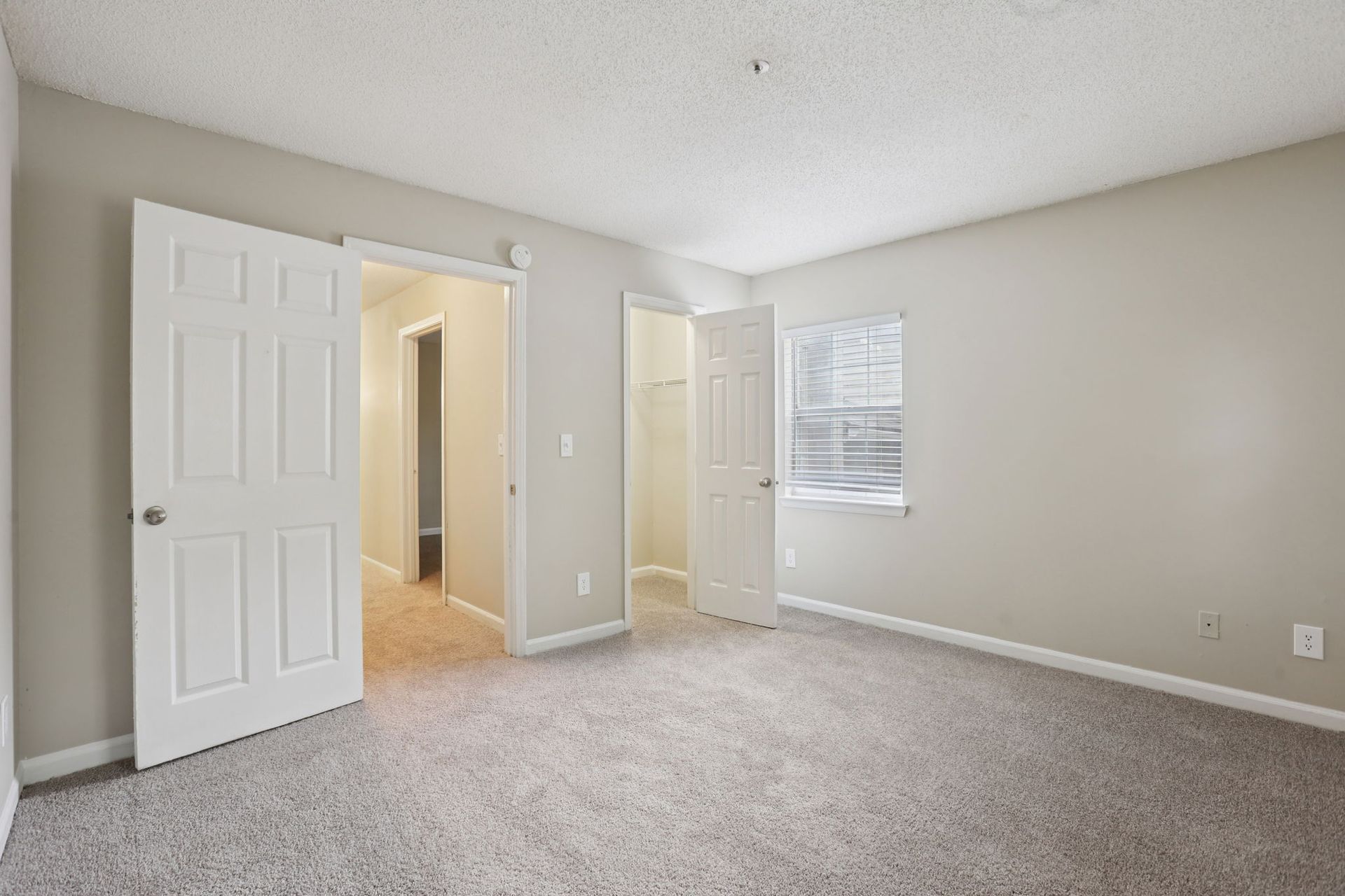 An empty room with light gray carpet, beige walls, a window, and two white doors leading to closets or hallways.