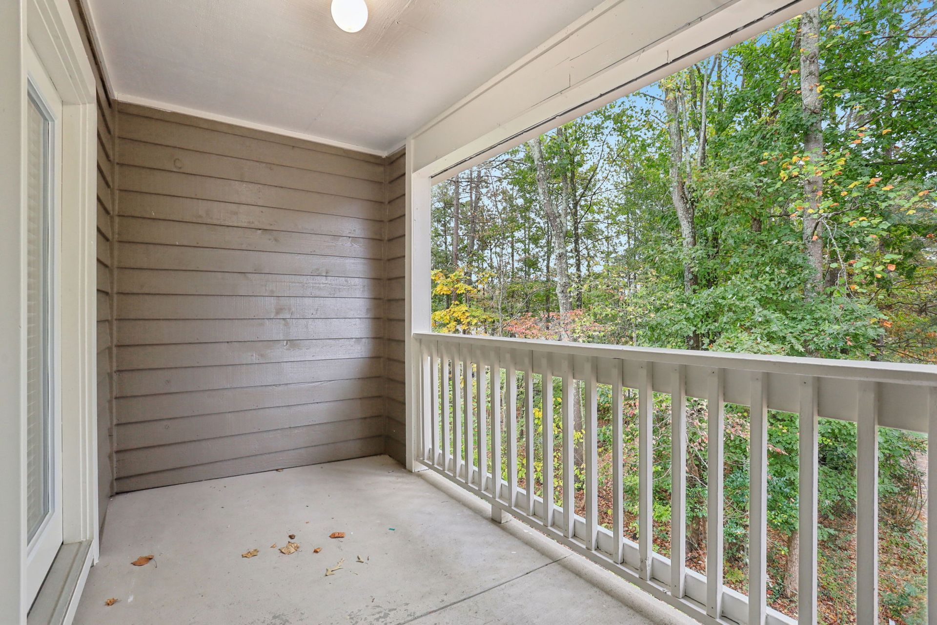 An outdoor balcony with beige concrete flooring, brown horizontal wood-paneled walls, and white railings facing a forest.