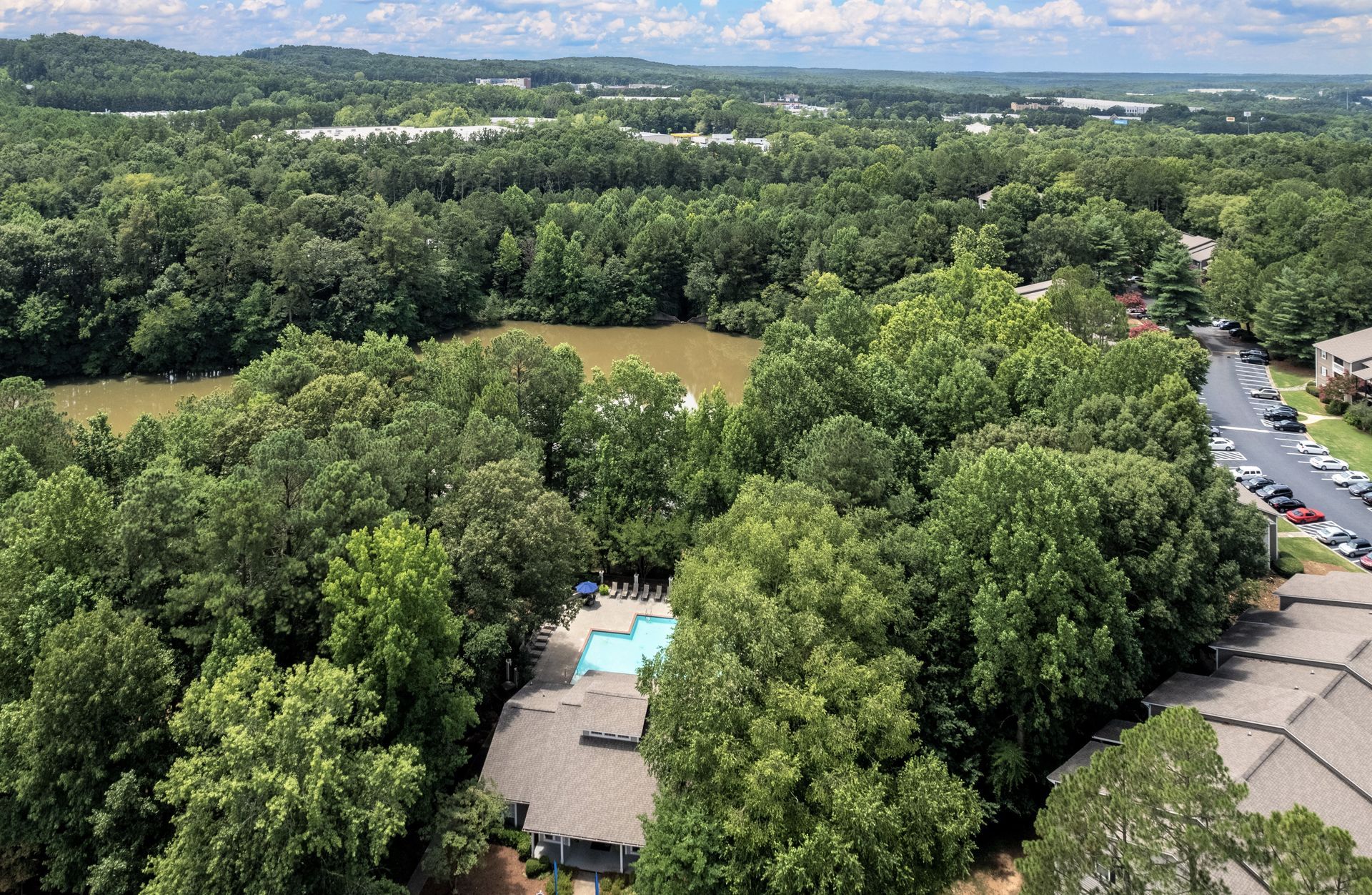 An aerial view of an apartment complex, showing a swimming pool, wooded area, pond, and parking lot under a cloudy sky.