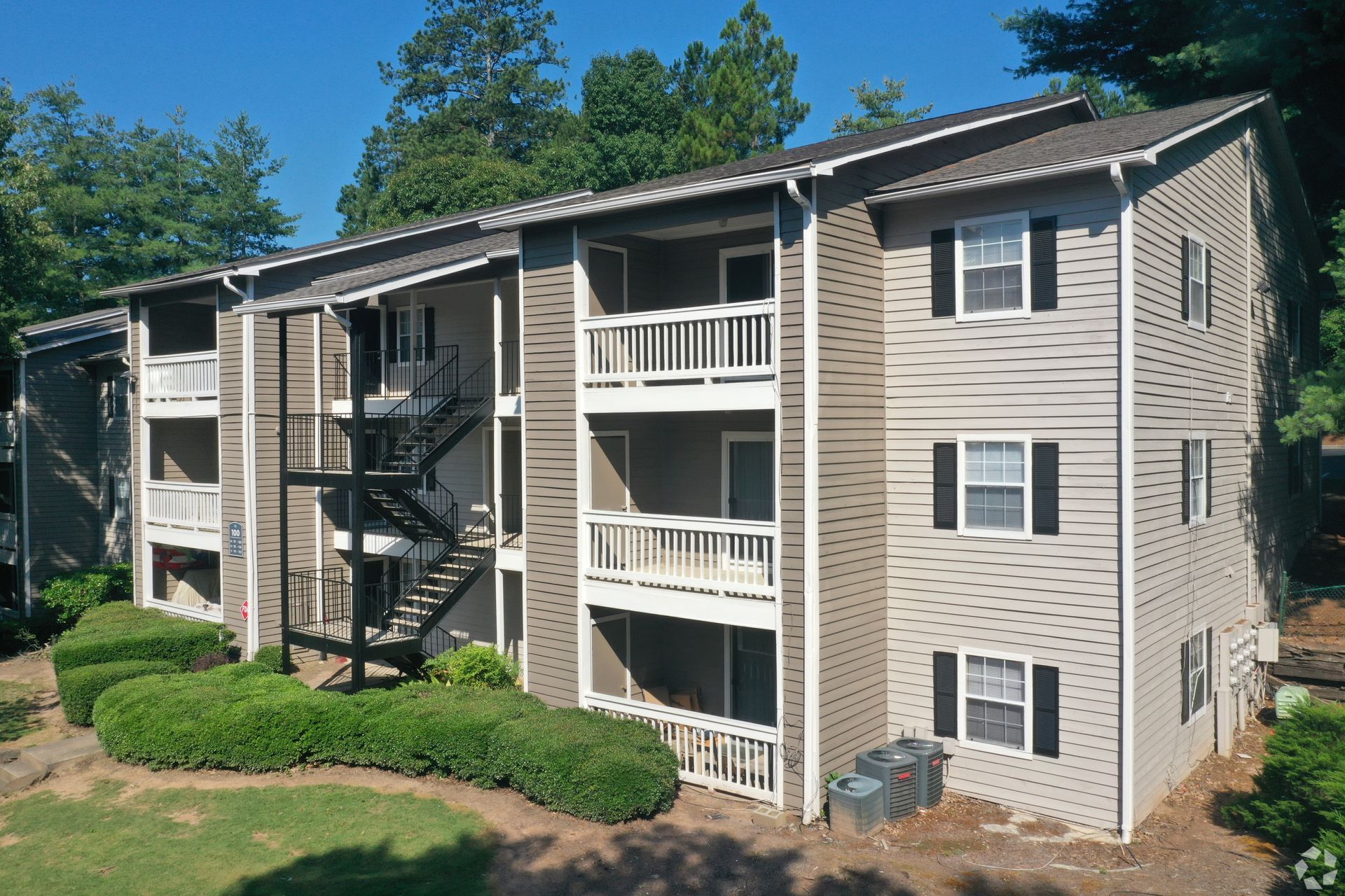 A three-story apartment building with beige siding, dark shutters, and outdoor staircases surrounded by trees and shrubs.